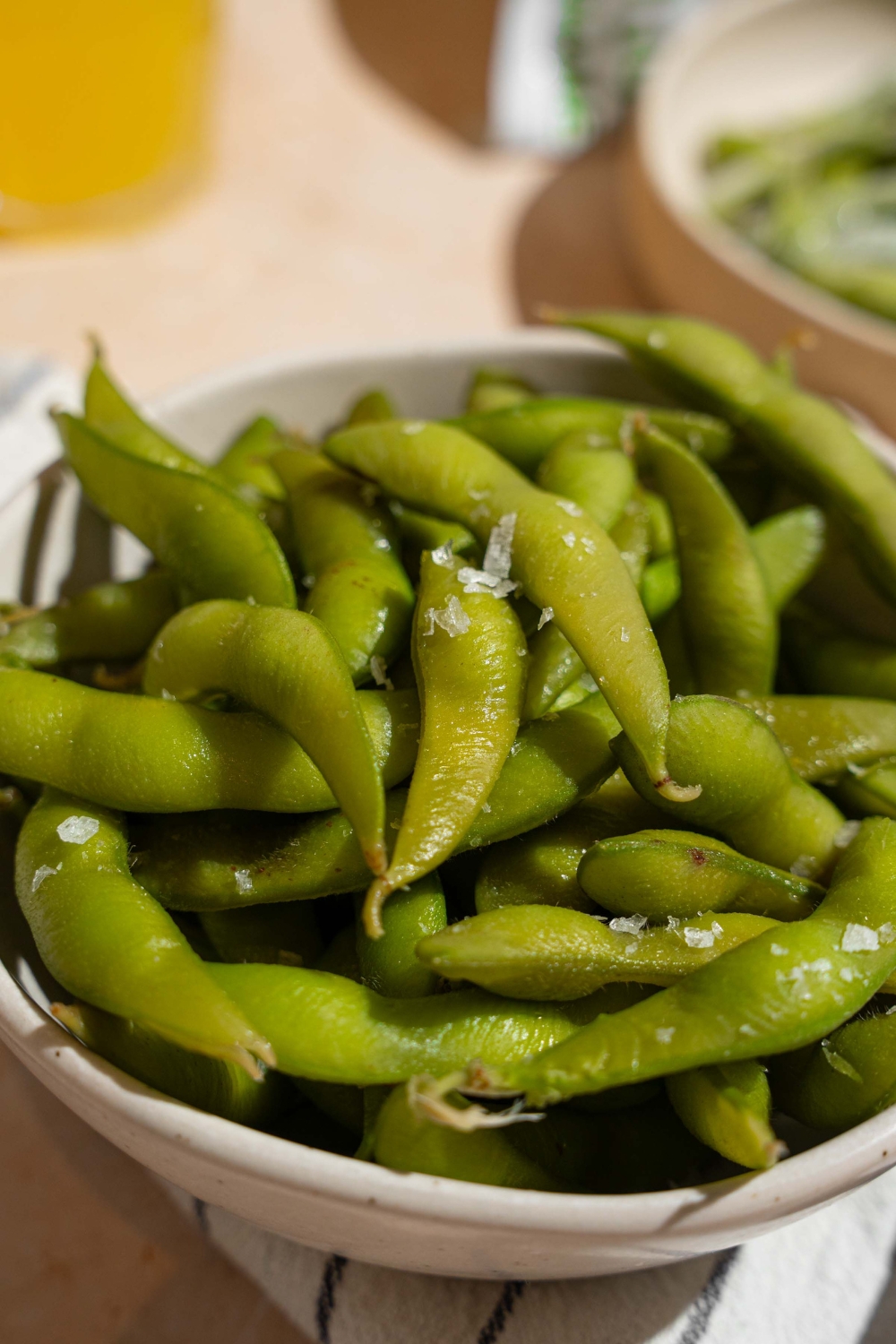 A bowl of cooked edamame seasoned with salt. The bowl is on a white striped napkin on a tan counter with a plate of frozen edamame.