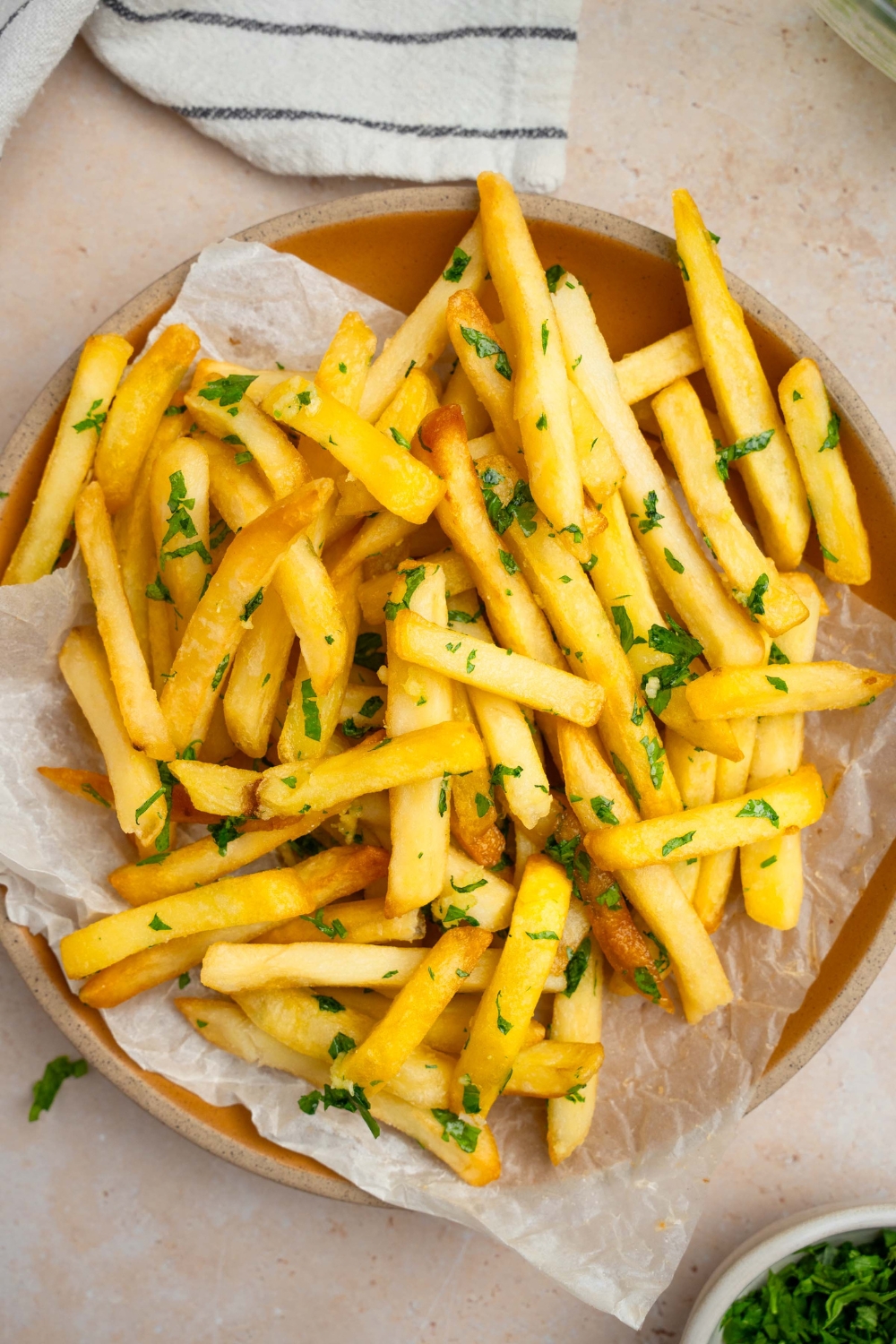 Garlic fries garnished with fresh parsley served on a plate lined with parchment paper. The plate is on a tan counter with a white striped napkin.