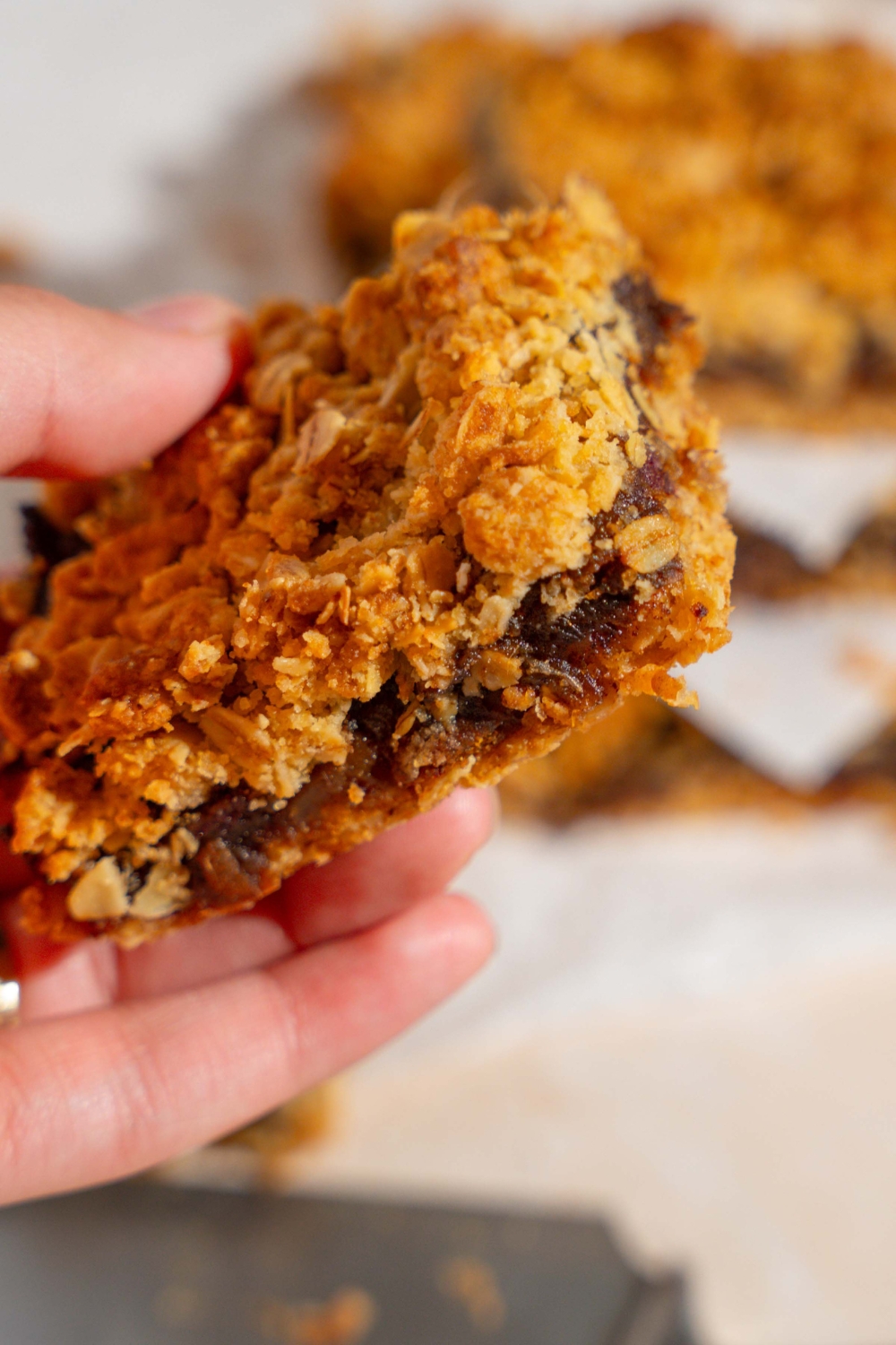 A close up of a hand holding an old fashioned date bar. There is a board of bars lined with parchment paper blurred in the background.
