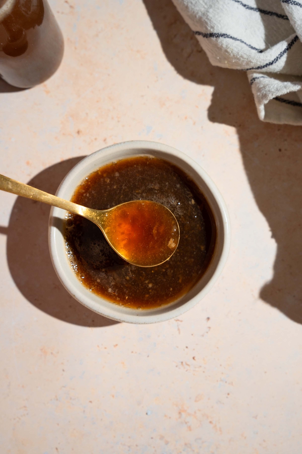 A bowl of oyster sauce with a spoon. The bowl is on a tan counter with a white striped napkin and jar of sauce.
