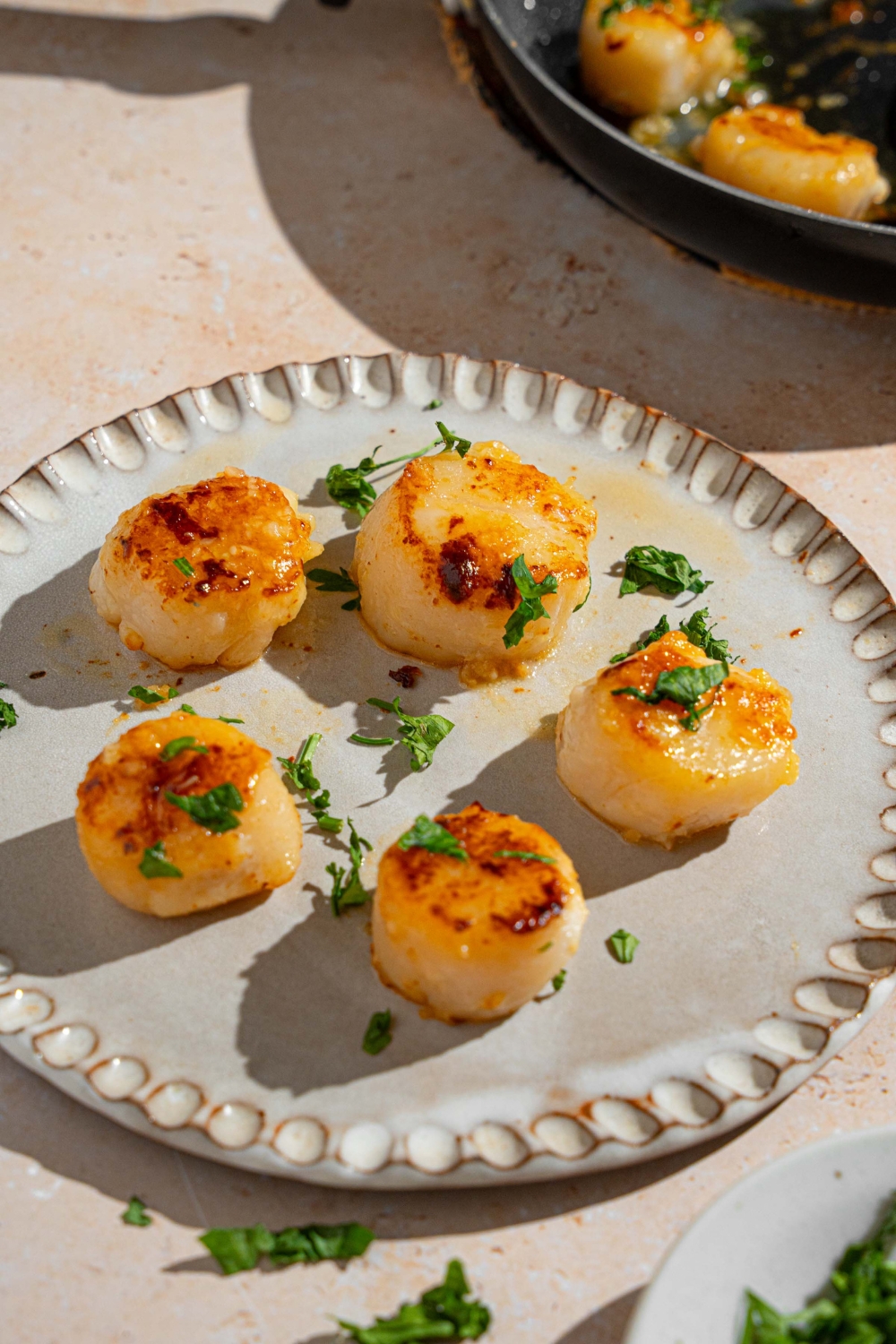 A ceramic plate with several pan seared scallops garnished with fresh parsley. The plate is on a tan counter with a skillet of scallops.