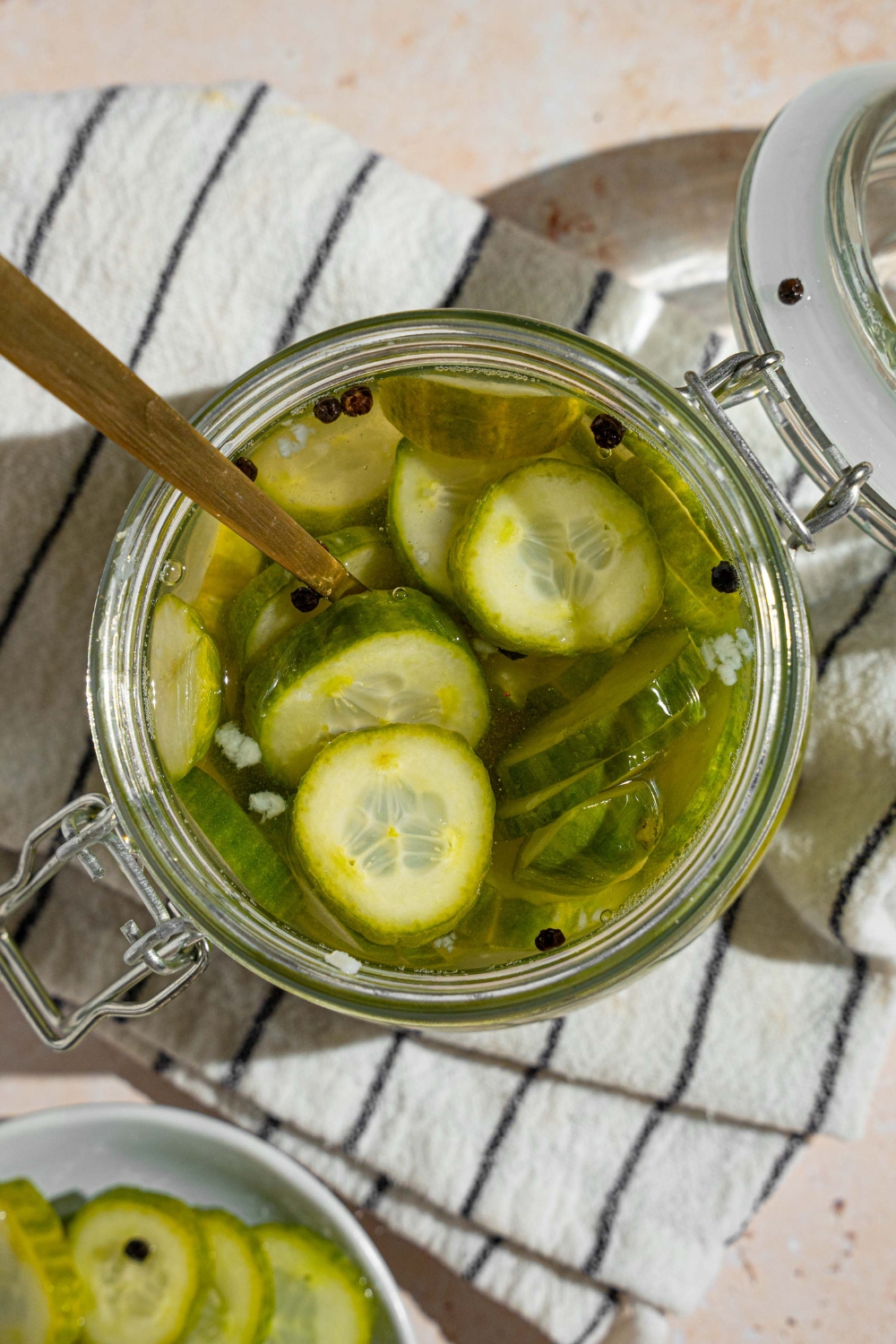A jar with quick pickles in juices. There is a fork in the jar. The jar is on a white striped napkin on a tan counter with a small plate of sliced pickles.