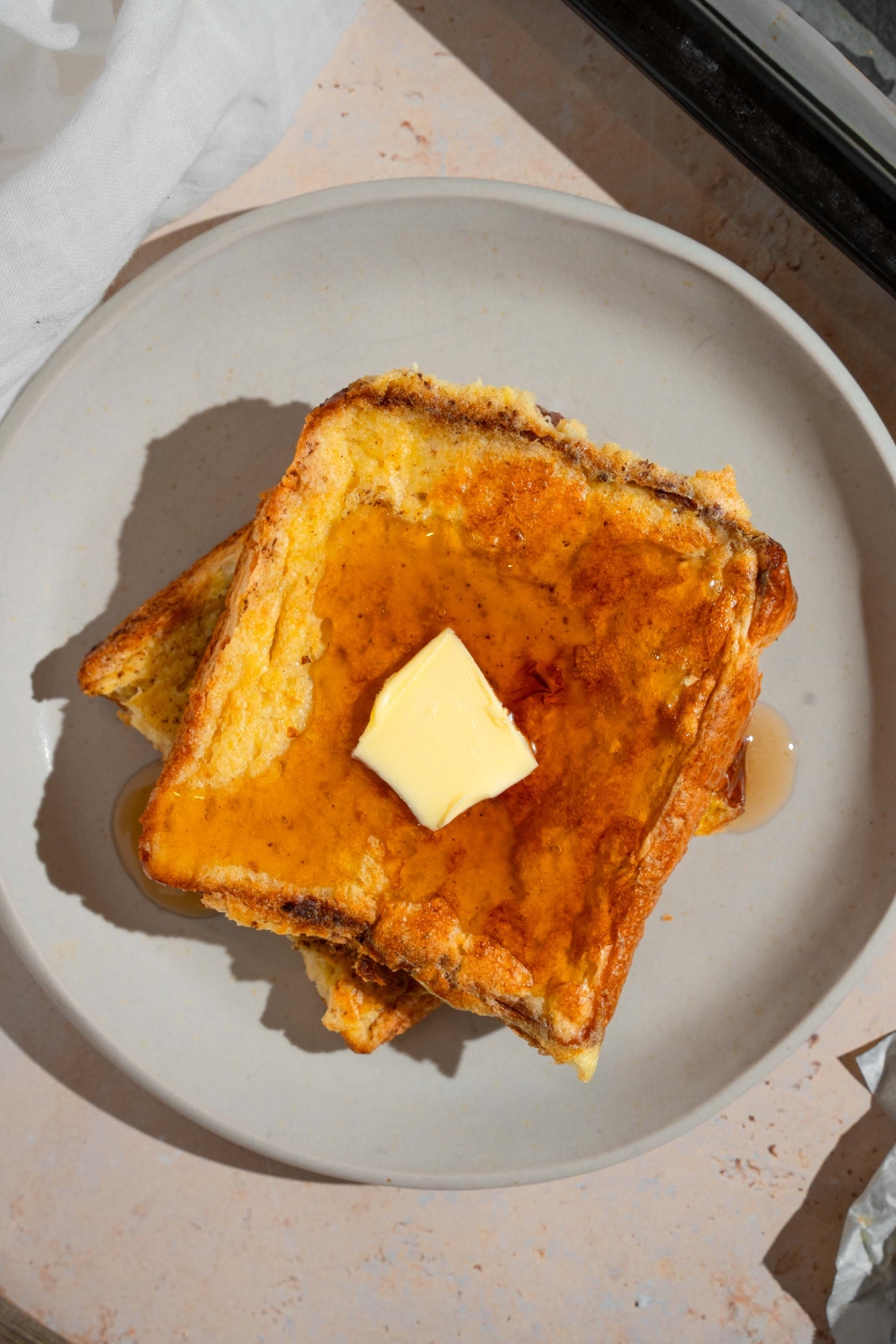 A plate with two slices of sheet pan french toast topped with butter and drizzled with syrup. The plate is on a tan counter.