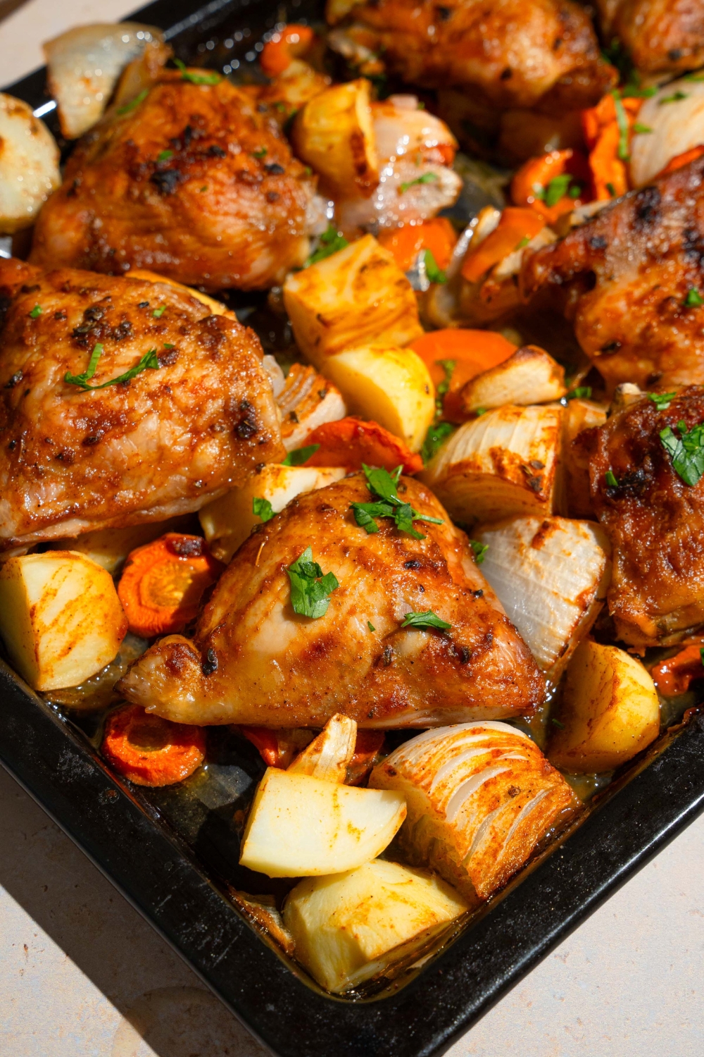 A baking sheet with roasted chicken thighs, carrots, potatoes, and onions garnished with fresh parsley.