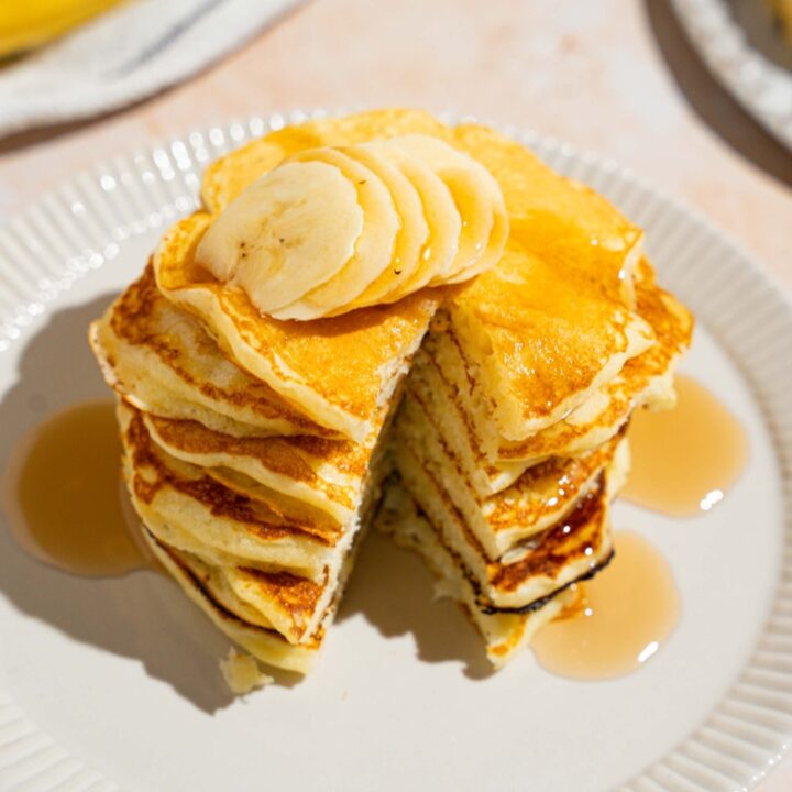 A stack of pancakes topped with sliced banana and drizzled with syrup. The pancakes are on a white plate and there is a bite removed from the stack. The plate is on a tan counter with a white striped napkin and bananas.