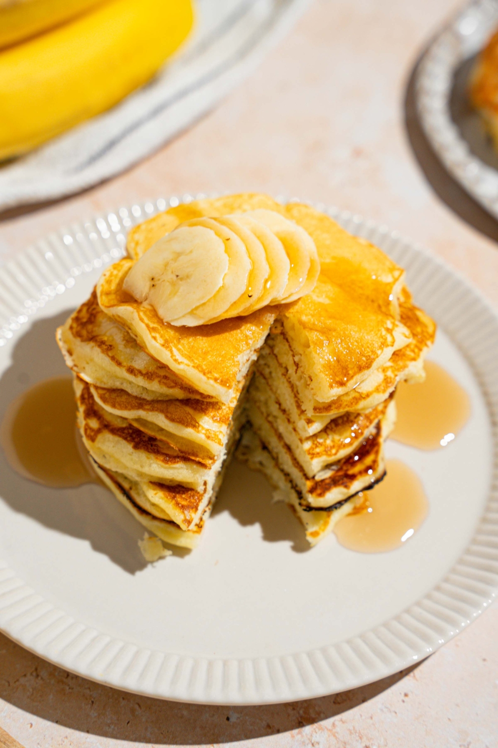 A stack of pancakes topped with butter and drizzled with syrup. The pancakes are on a white plate and there is a bite removed from the stack. The plate is on a tan counter with a white striped napkin and bananas.
