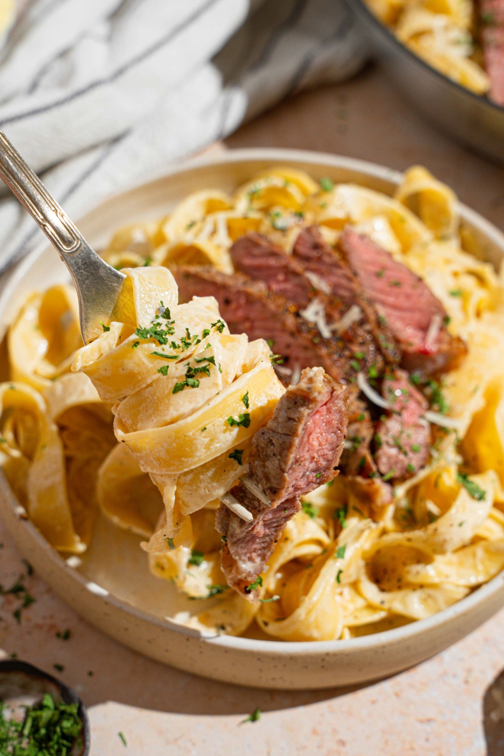 Fettuccine alfredo topped with sliced steak garnished with fresh parsley and grated cheese in a white bowl. There is a fork twirling a bite of pasta. The bowl is on a tan counter with a fork and white striped napkin.