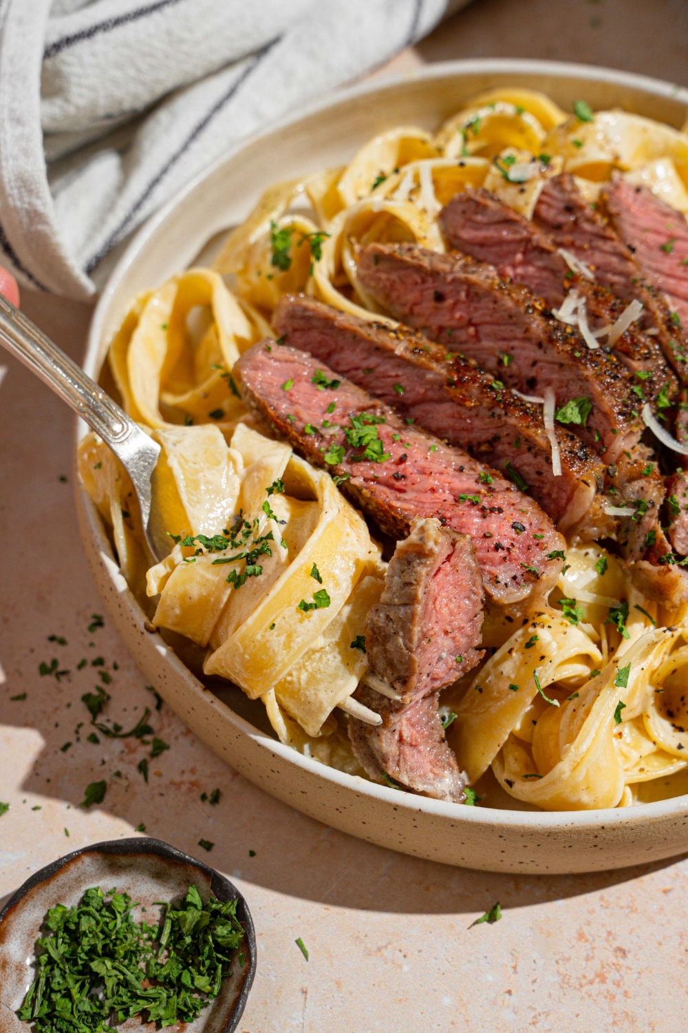 Fettuccine alfredo topped with sliced steak garnished with fresh parsley and grated cheese in a white bowl. There is a fork twirling a bite of pasta. The bowl is on a tan counter with a fork and white striped napkin.