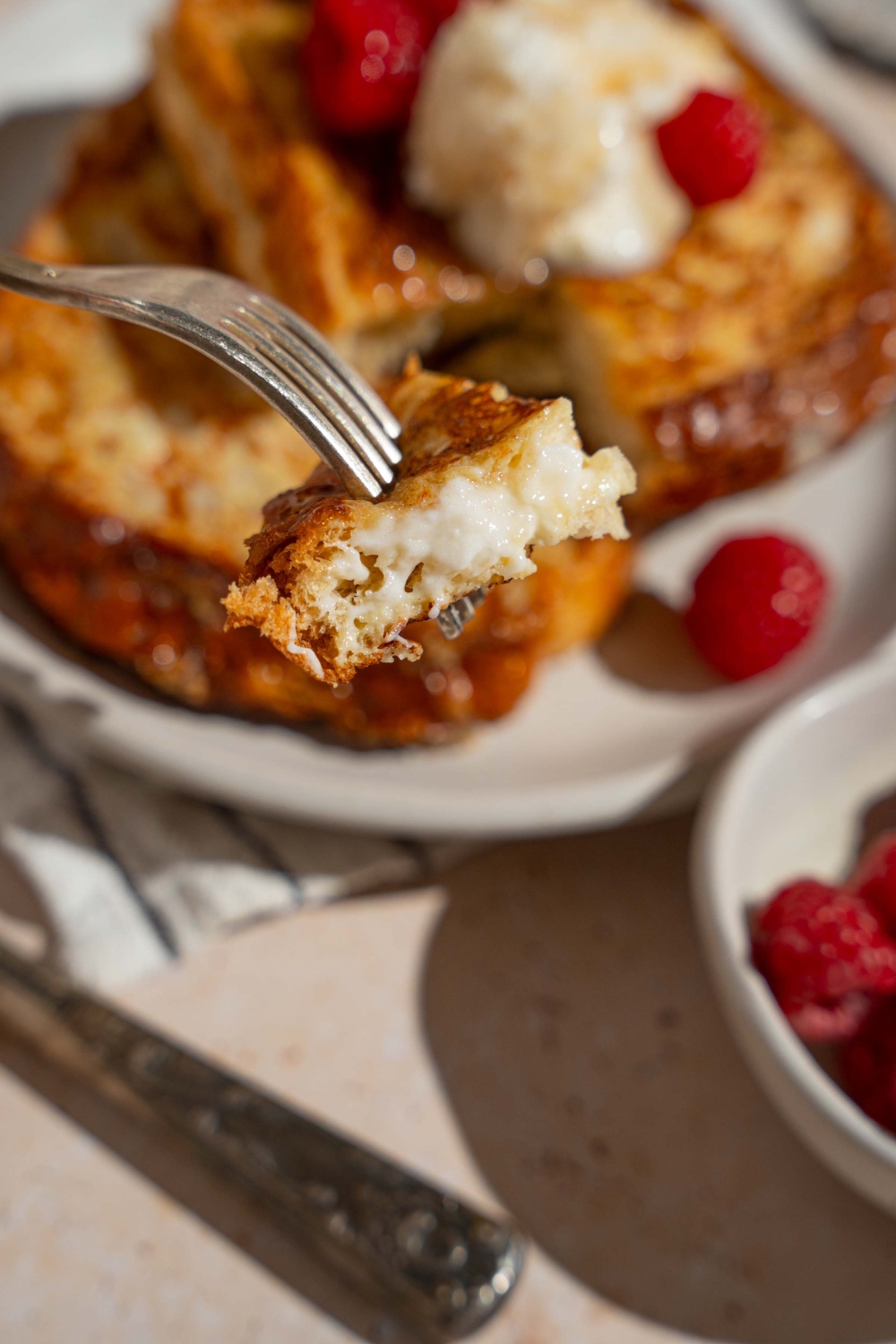 A close up of a fork with a bite of stuffed french toast. There is a plate of french toast with raspberries blurred in the background.