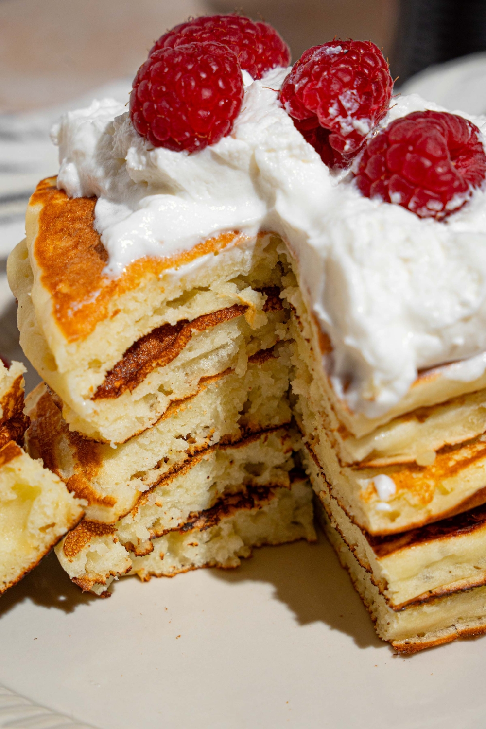 A white plate with a stack of sweet cream fluffy pancakes topped with whipped cream and raspberries. A bite has been taken from the stack.