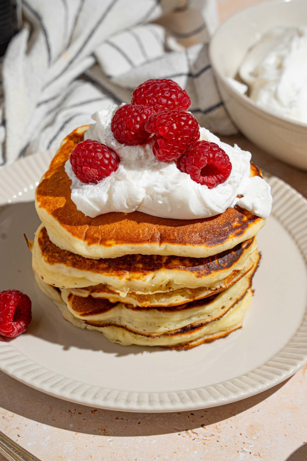 A white plate with a stack of sweet cream pancakes topped with whipped cream and raspberries. The plate is on a tan counter with a white striped napkin and bowl of whipped cream.