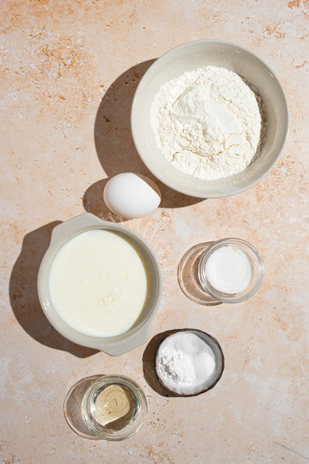 An overhead shot of several bowls in various sizes containing ingredients to make small batch pancakes including flour, buttermilk, egg, oil, baking soda, baking powder, and salt.