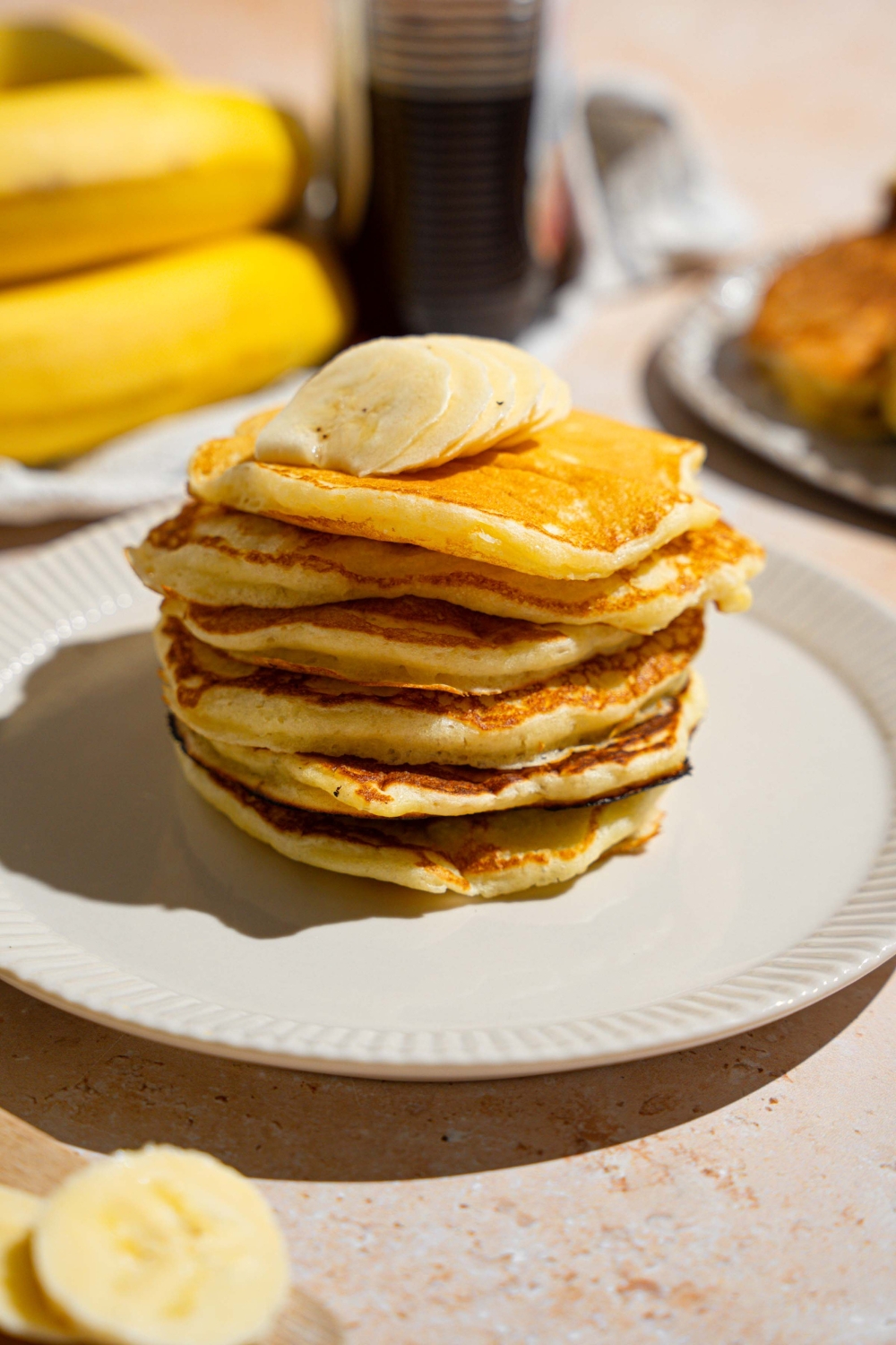 A stack of pancakes topped with sliced banana. The pancakes are on a white plate. The plate is on a tan counter with a white striped napkin and bananas.
