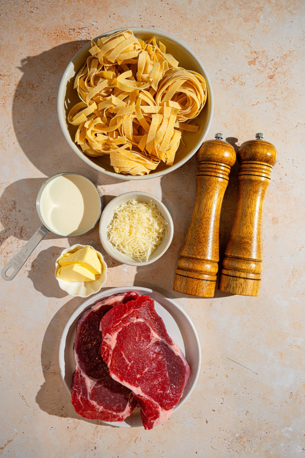 An overhead shot of several bowls in various sizes containing ingredients to make steak alfredo including steaks, fettuccine pasta, grated cheese, butter, cream, and a salt and pepper mill.
