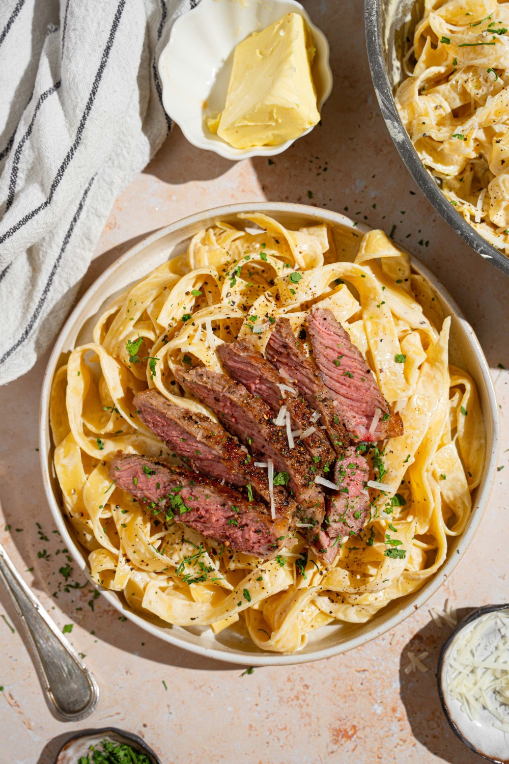 Fettuccine alfredo topped with sliced steak garnished with fresh parsley and grated cheese in a white bowl. There is a fork twirling a bite of pasta. The bowl is on a tan counter with a fork and white striped napkin.