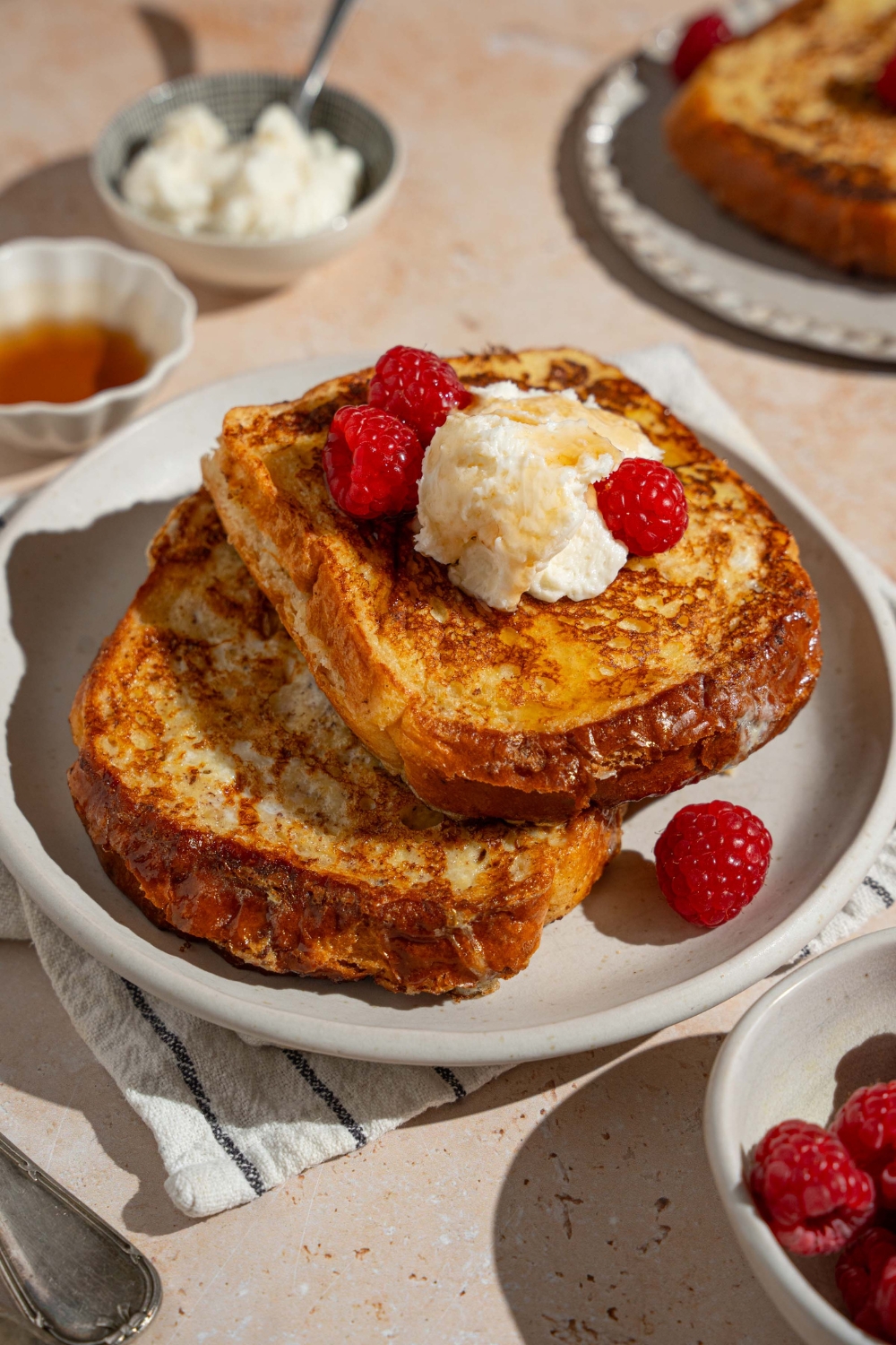 A white plate with stuffed french toast topped with whipped cream cheese and raspberries drizzled with syrup. The plate is on a tan counter with a white striped napkin and bowl of raspberries.