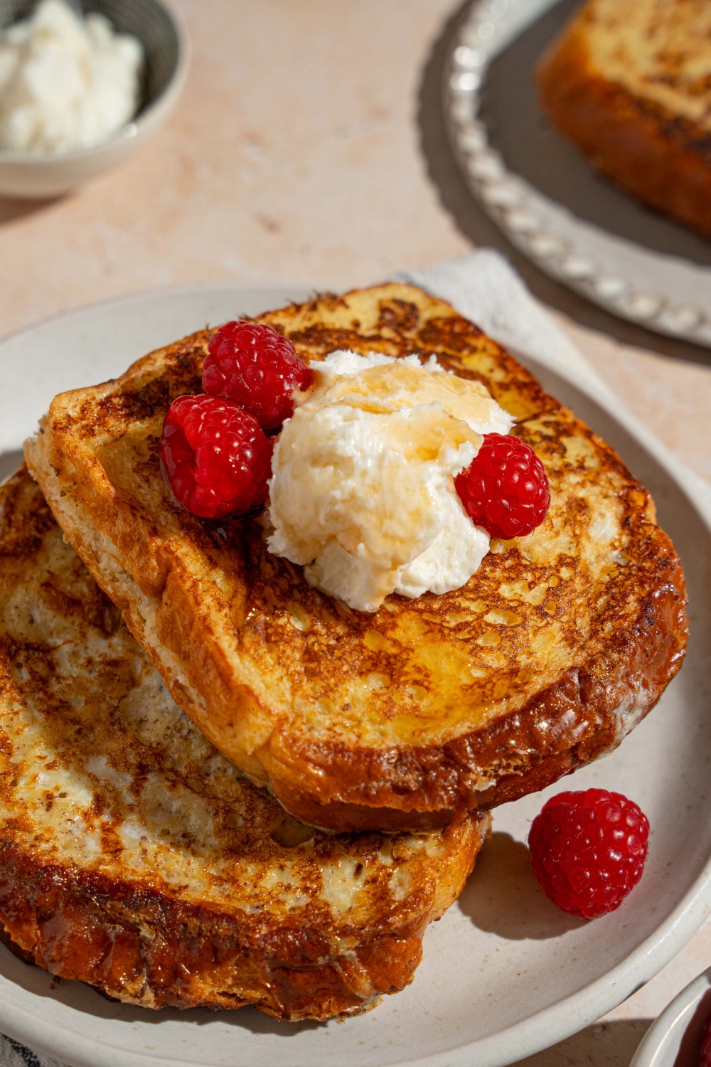 A white plate with stuffed french toast topped with whipped cream cheese and raspberries drizzled with syrup. The plate is on a tan counter with a white striped napkin and bowl of cream cheese.