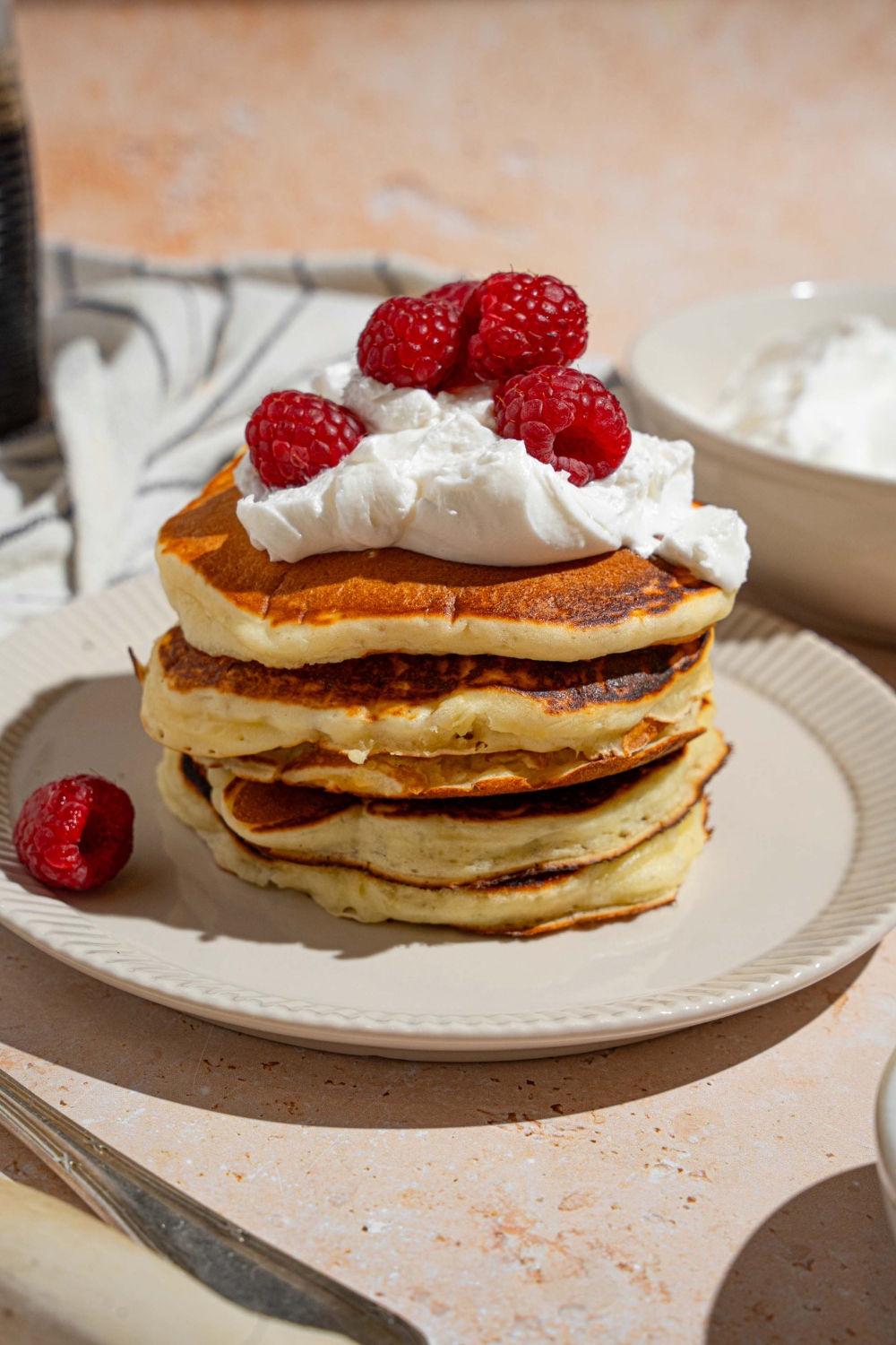 A white plate with a stack of sweet cream pancakes topped with whipped cream and raspberries. The plate is on a tan counter with a white striped napkin and bowl of whipped cream.