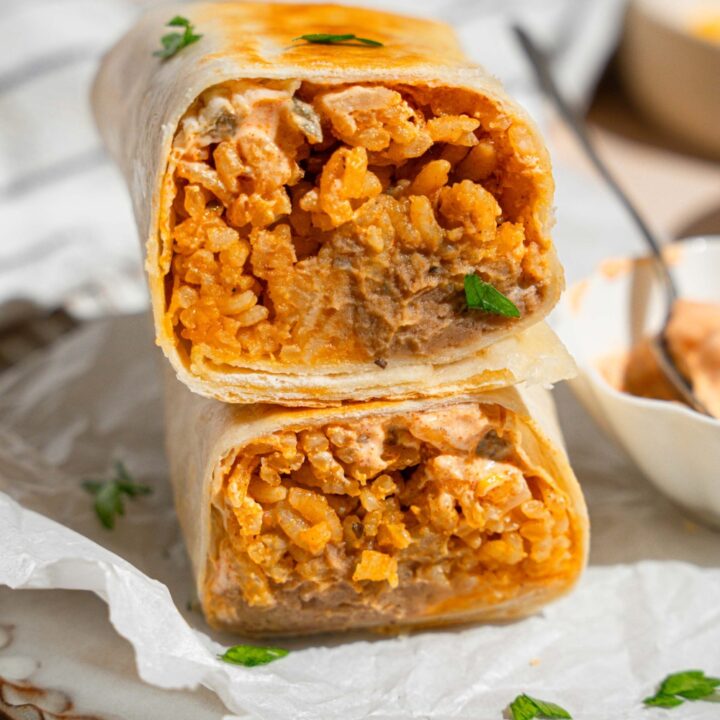 A cheesy bean and rice burrito sliced in half with each half stacked on the other. The burrito is on a plate lined with parchment paper with a small side of creamy jalapeno sauce with a spoon. The plate is garnished with fresh parsley. The plate is on a tan counter.