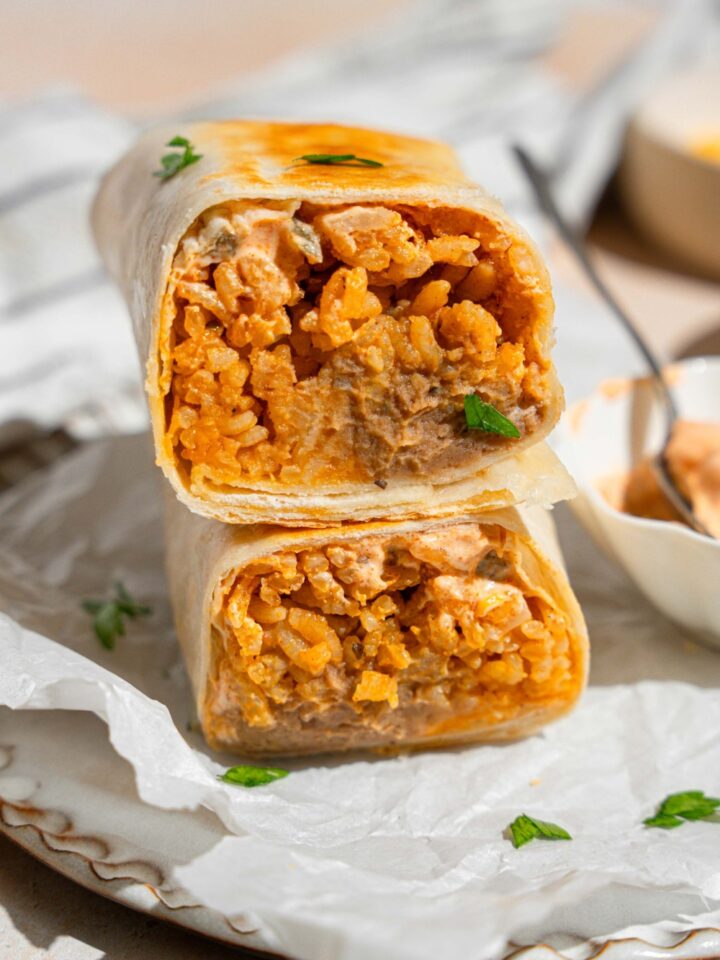 A cheesy bean and rice burrito sliced in half with each half stacked on the other. The burrito is on a plate lined with parchment paper with a small side of creamy jalapeno sauce with a spoon. The plate is garnished with fresh parsley. The plate is on a tan counter.