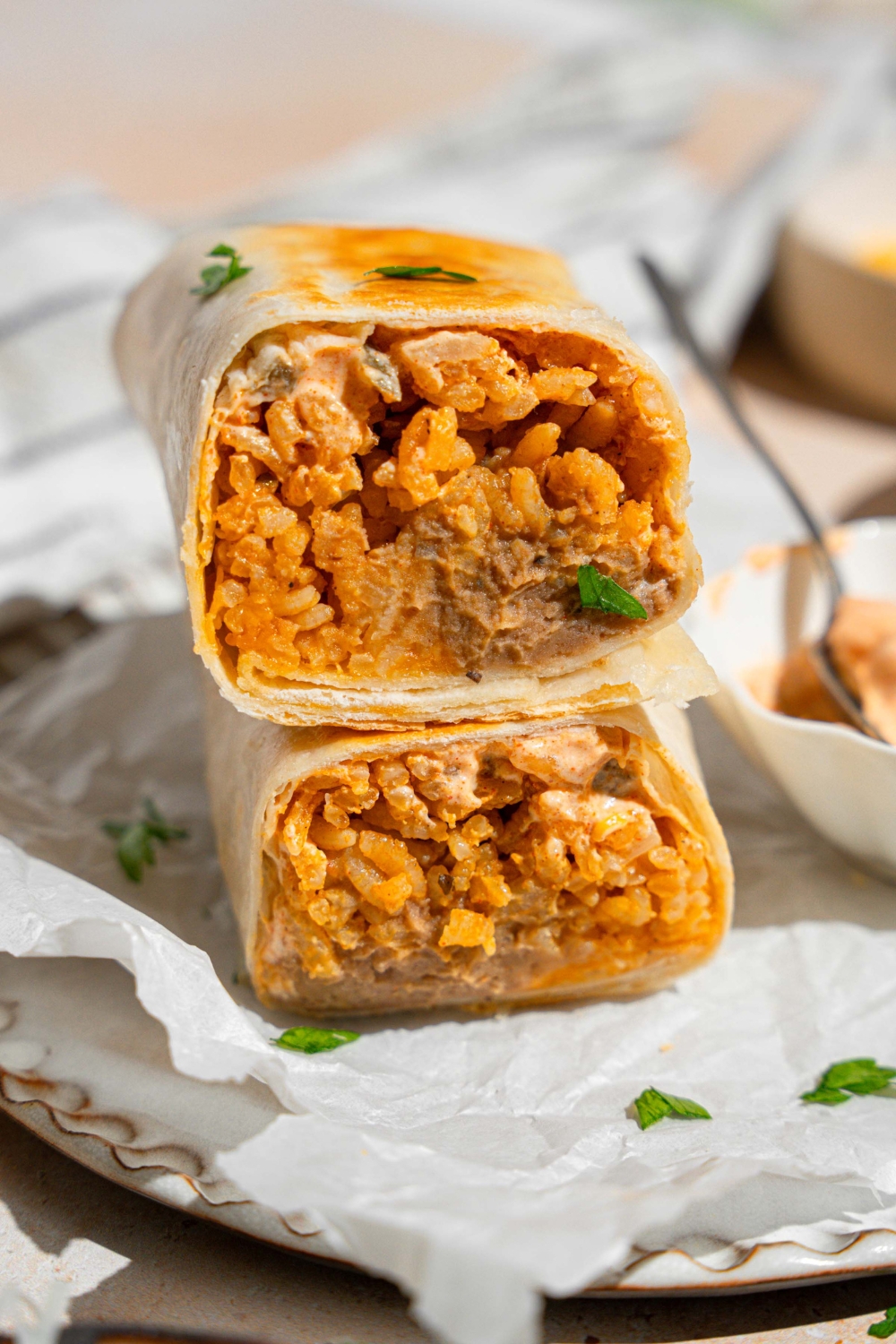 A cheesy bean and rice burrito sliced in half with each half stacked on the other. The burrito is on a plate lined with parchment paper with a small side of creamy jalapeno sauce with a spoon. The plate is garnished with fresh parsley. The plate is on a tan counter.