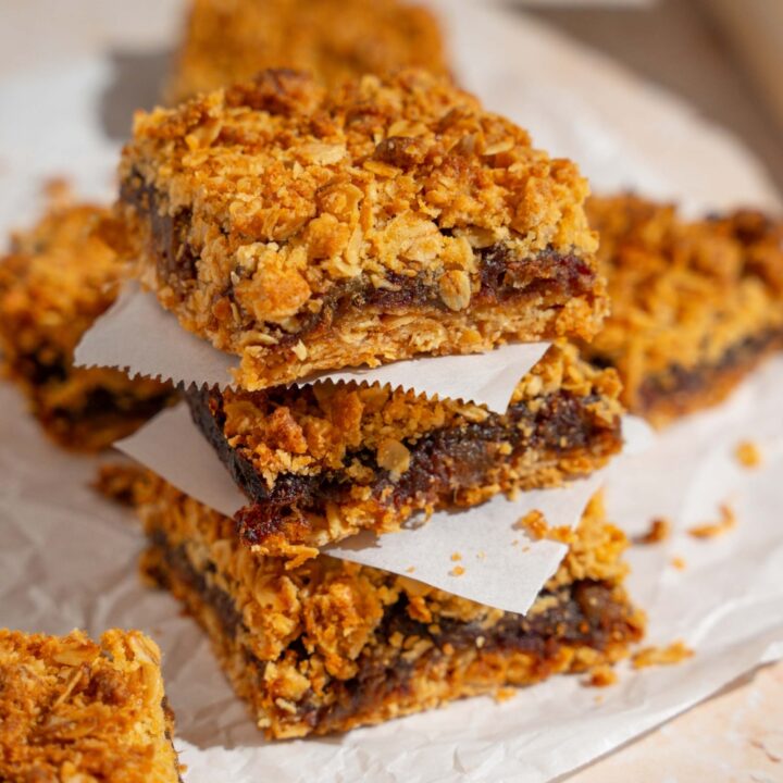 A stack of three old fashioned date bars with a parchment square in between each one. The stack is on a piece of parchment paper with additional bars.