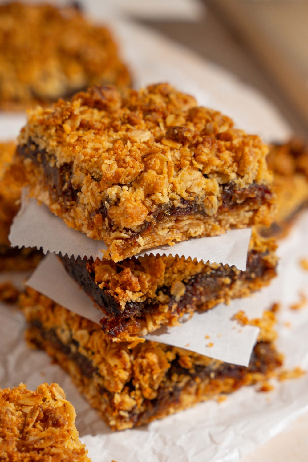 A stack of three old fashioned date bars with a parchment square in between each one. The stack is on a piece of parchment paper with additional bars.
