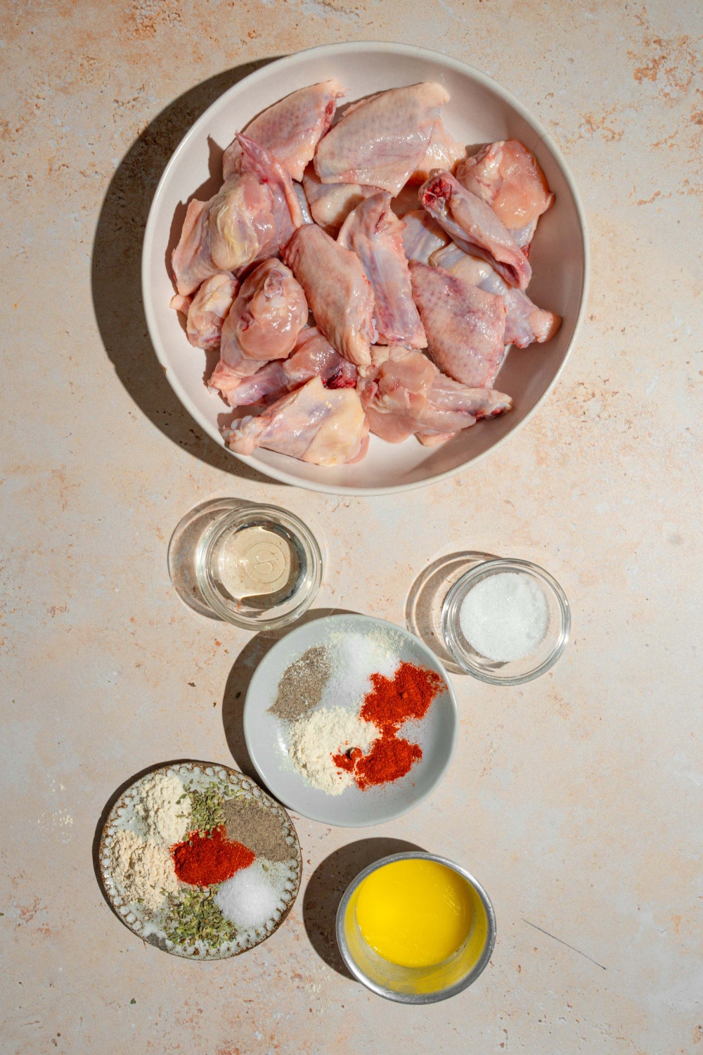 An overhead shot of several bowls in various sizes containing ingredients to make Wingstop Louisiana rub wings including wings, butter, oil, sugar, garlic powder, paprika, ground ginger, oregano, and more.