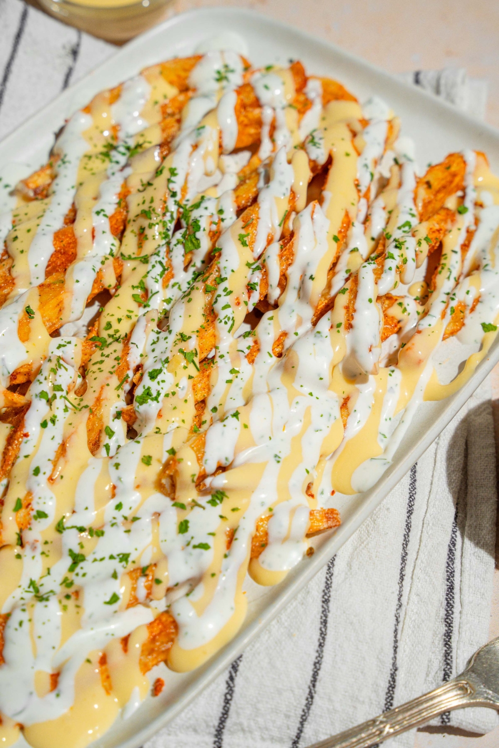 A white platter with copycat Wingstop Voodoo fries drizzled with cheese and ranch dressing. The plate is garnished with chopped parsley and served over a white striped napkin.