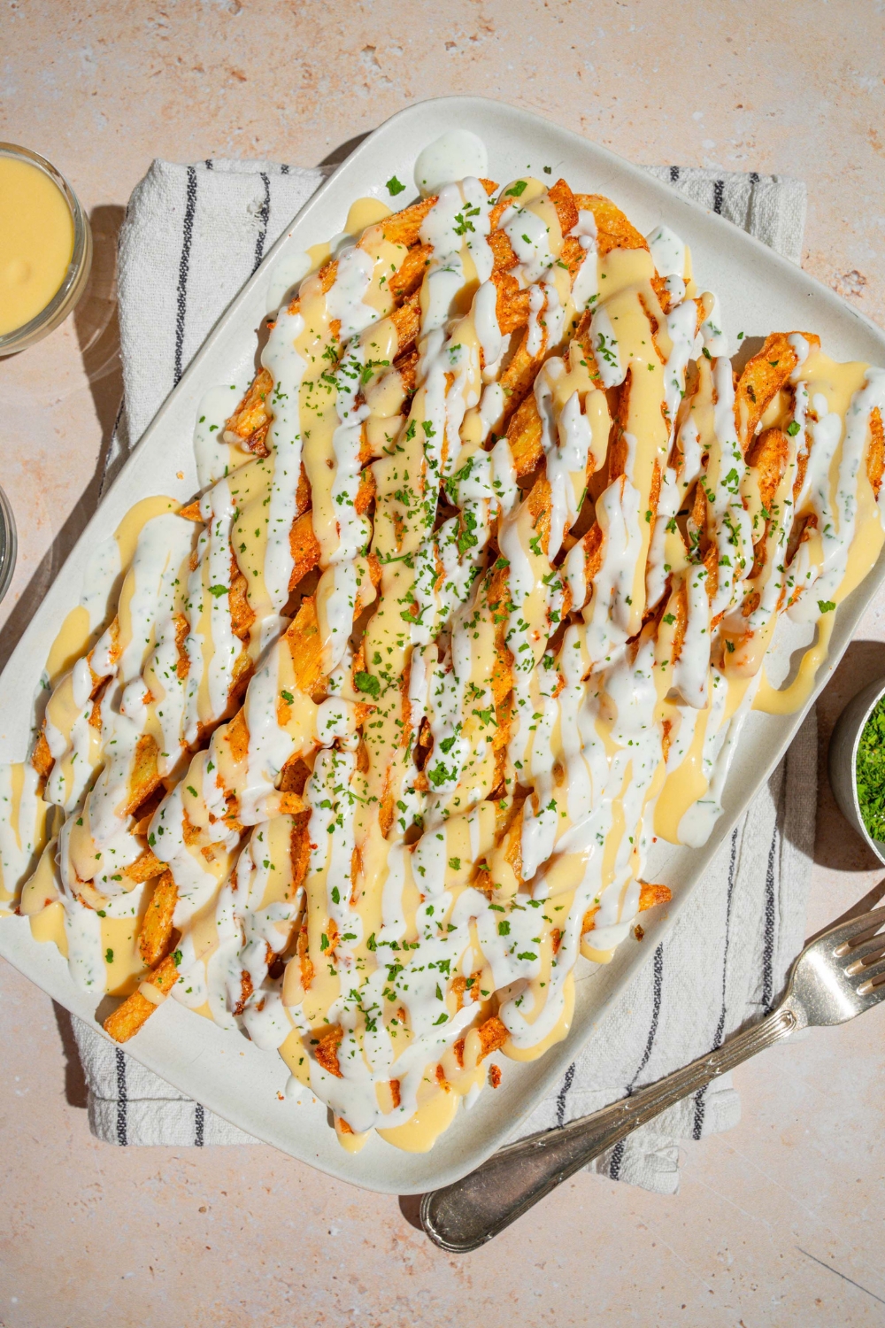 A white platter with copycat Wingstop Voodoo fries drizzled with cheese and ranch dressing. The plate is garnished with chopped parsley and served over a white striped napkin with a fork.