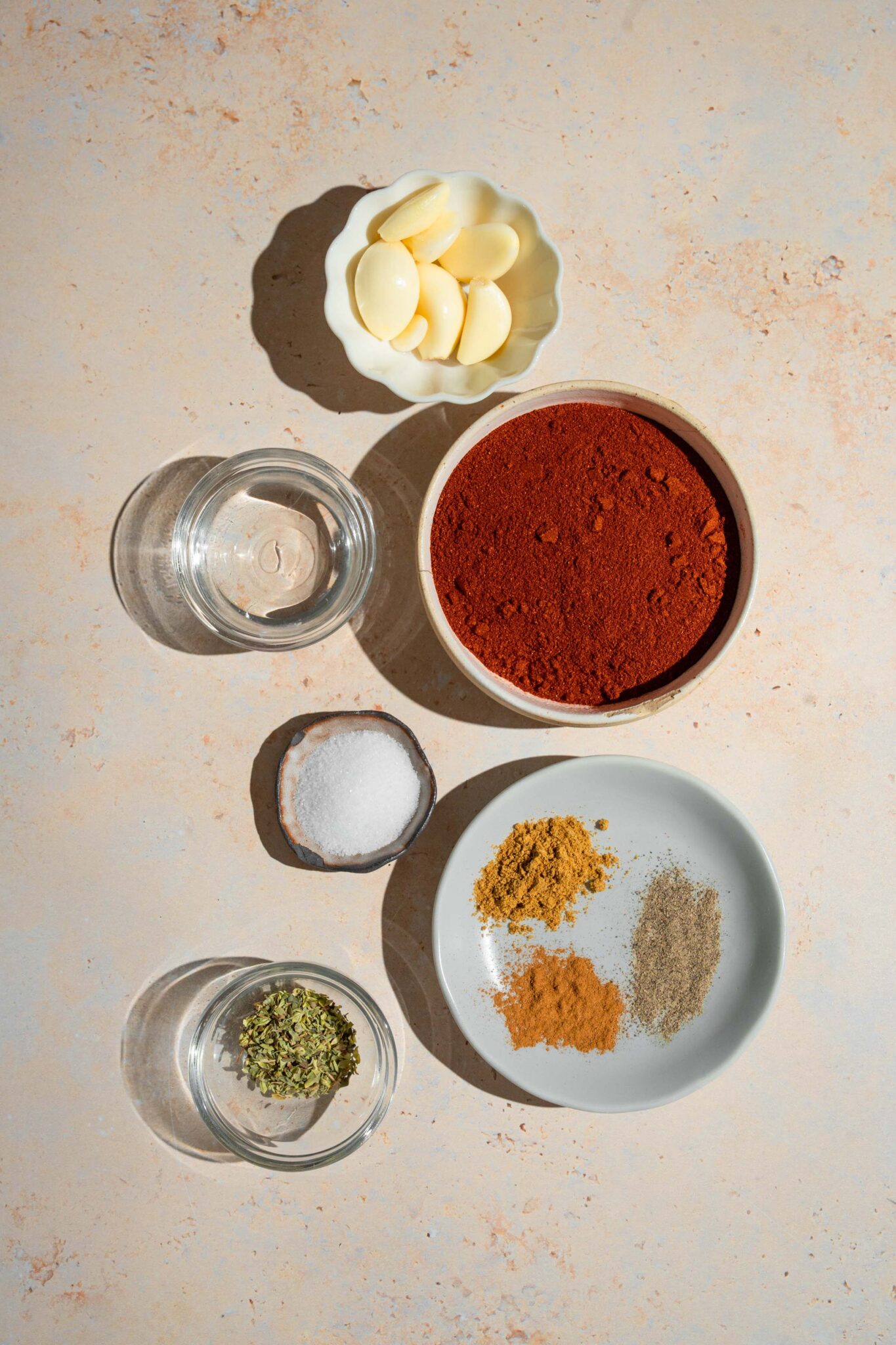 A bowl of garlic cloves, a bowl of adobo seasoning, a bowl of water, a bowl of salt, and a plate of seasonings all on a white counter.