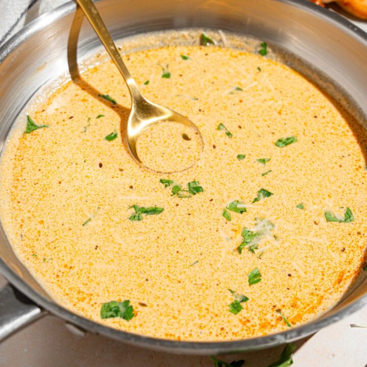 A skillet with a spoon stirring cajun alfredo sauce garnished with fresh parsley. The skillet is on a tan counter with a white striped napkin.