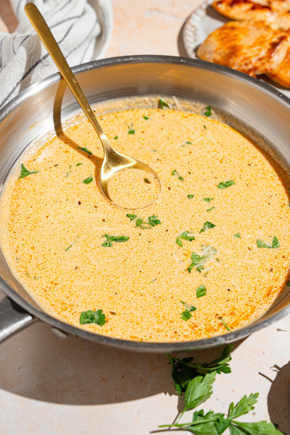 A skillet with a spoon stirring cajun alfredo sauce garnished with fresh parsley. The skillet is on a tan counter with a white striped napkin.