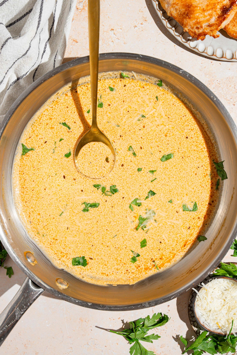 A skillet with a spoon stirring cajun alfredo sauce garnished with fresh parsley. The skillet is on a tan counter with a white striped napkin and plate of chicken.