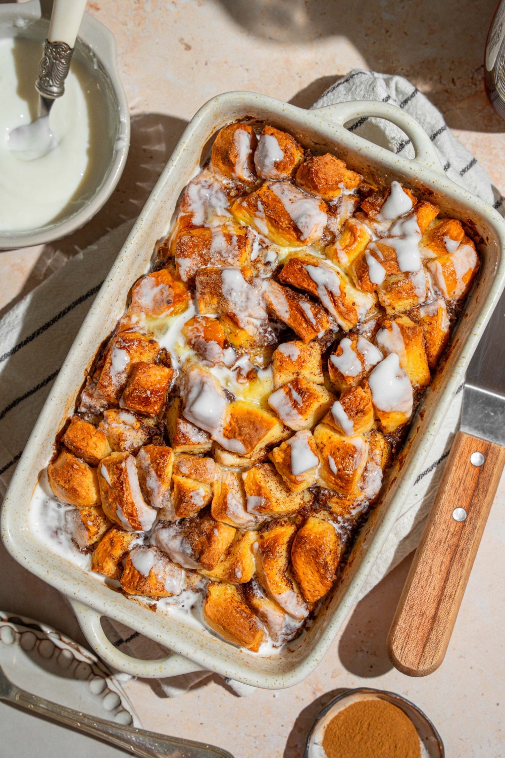 A baking dish with cinnamon roll french toast drizzled with icing. The dish is on a tan counter on a white striped napkin. The dish is on a tan counter with a small bowl of icing.