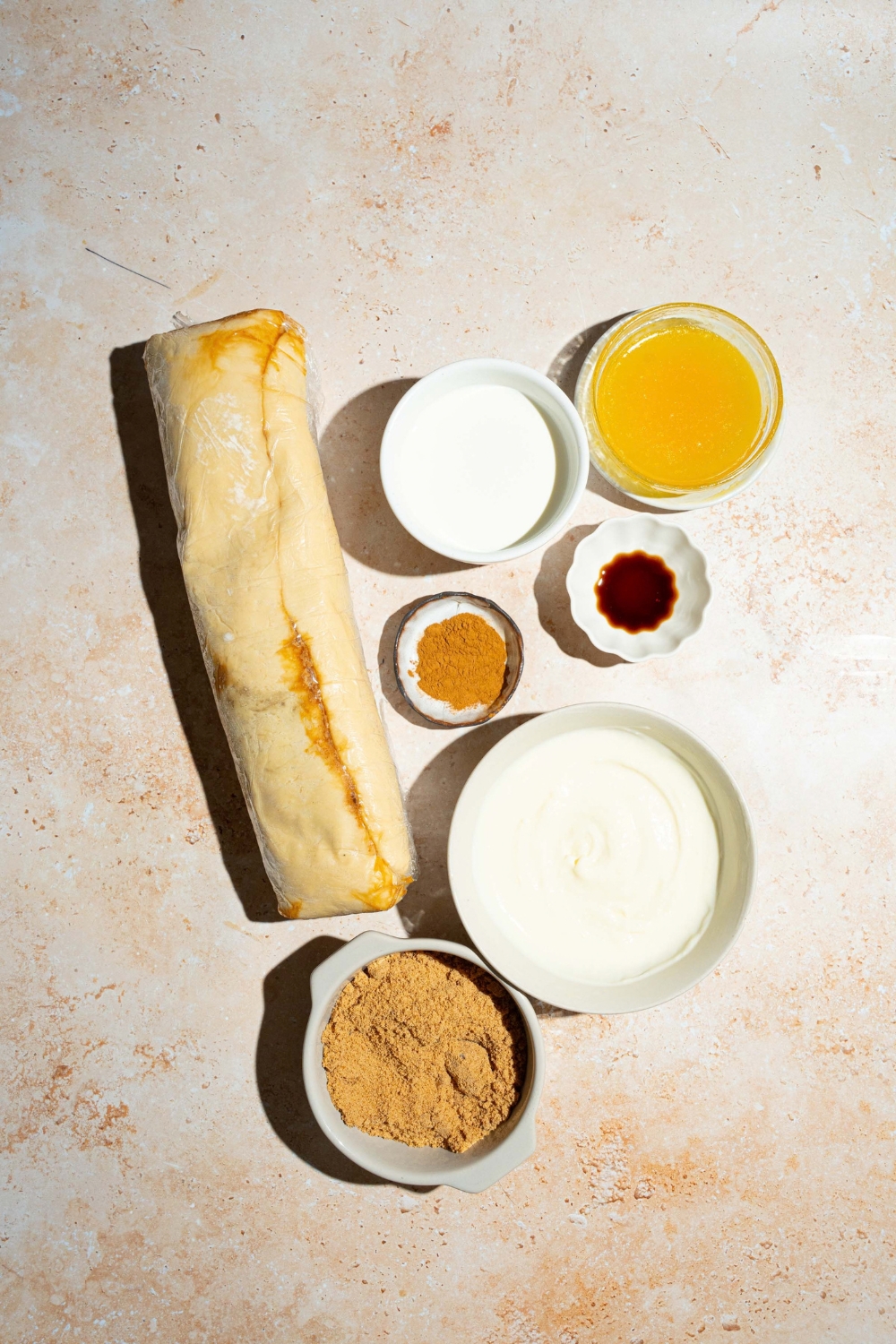 A package of cinnamon rolls, a bowl of heavy cream, a bowl of melted butter, a bowl of vanilla, a bowl of cinnamon, and a bowl of brown sugar.