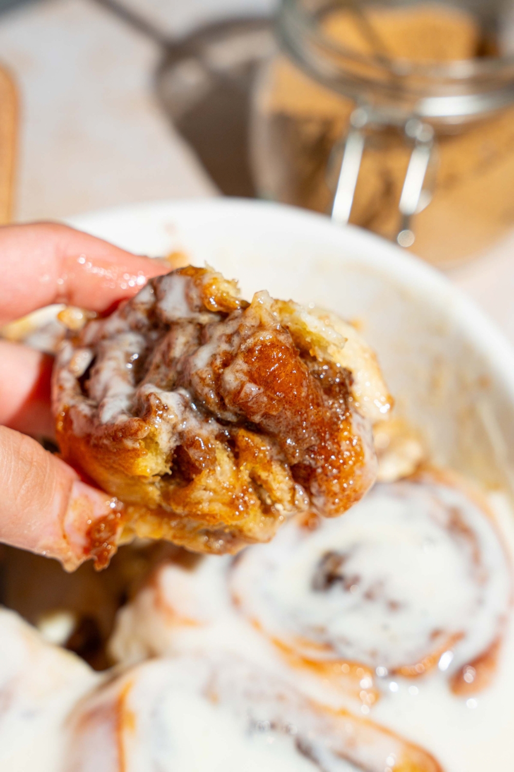 A hand holding a heavy cream cinnamon roll above a baking dish filled with the cinnamon rolls.