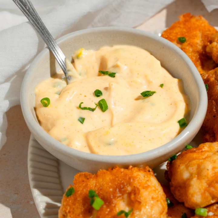 A white bowl with remoulade sauce in it and a spoon in the sauce. The bowl is on a plate with some fried shrimp on it.