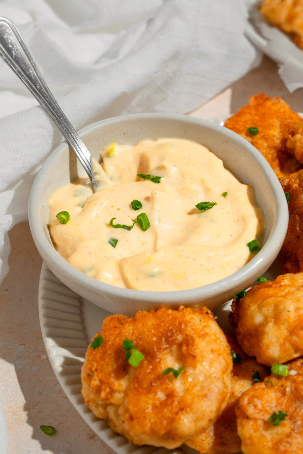 A white bowl with remoulade sauce in it and a spoon in the sauce. The bowl is on a plate with some fried shrimp on it.