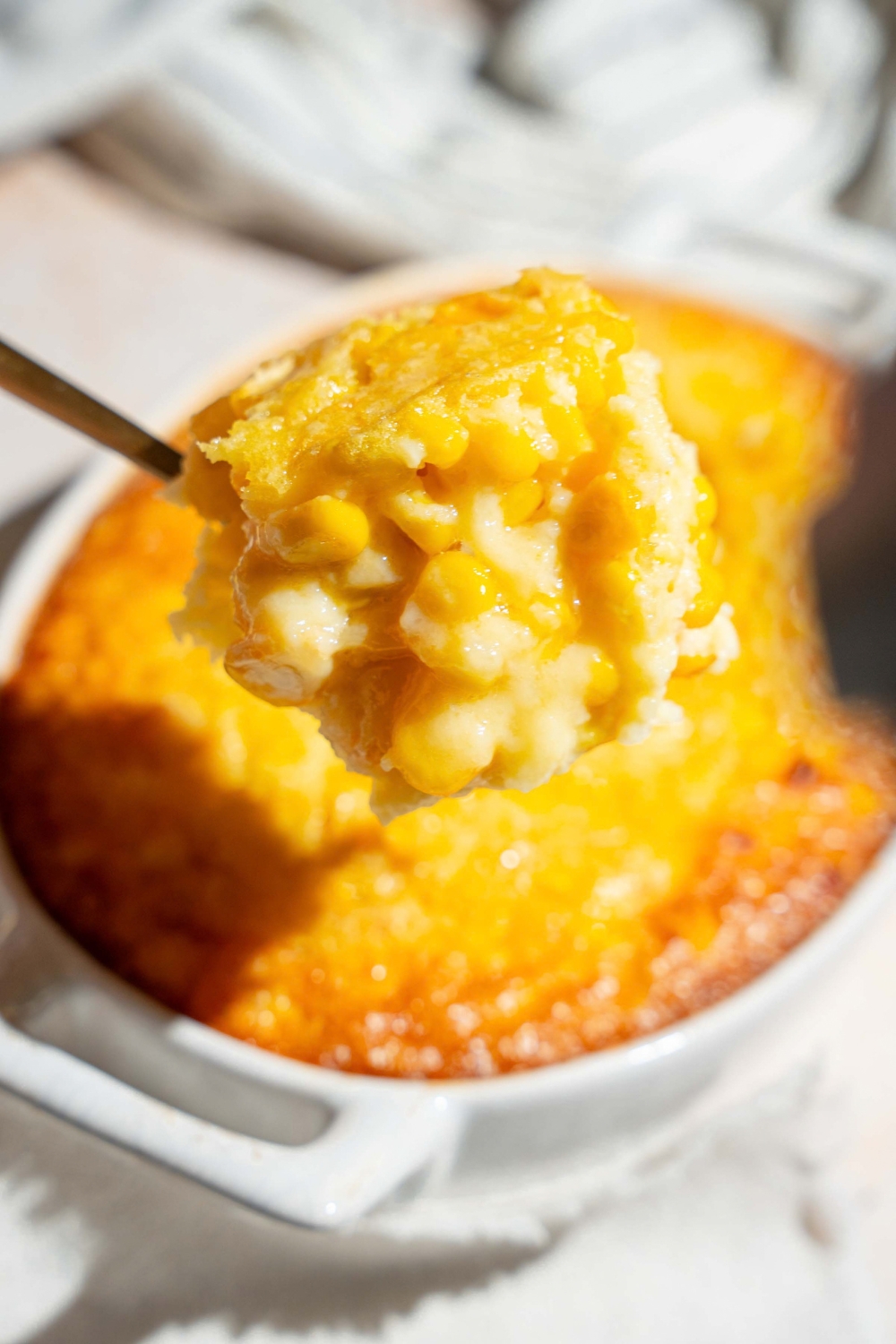 a scoop of corn casserole being held above a casserole dish with the corn casserole.