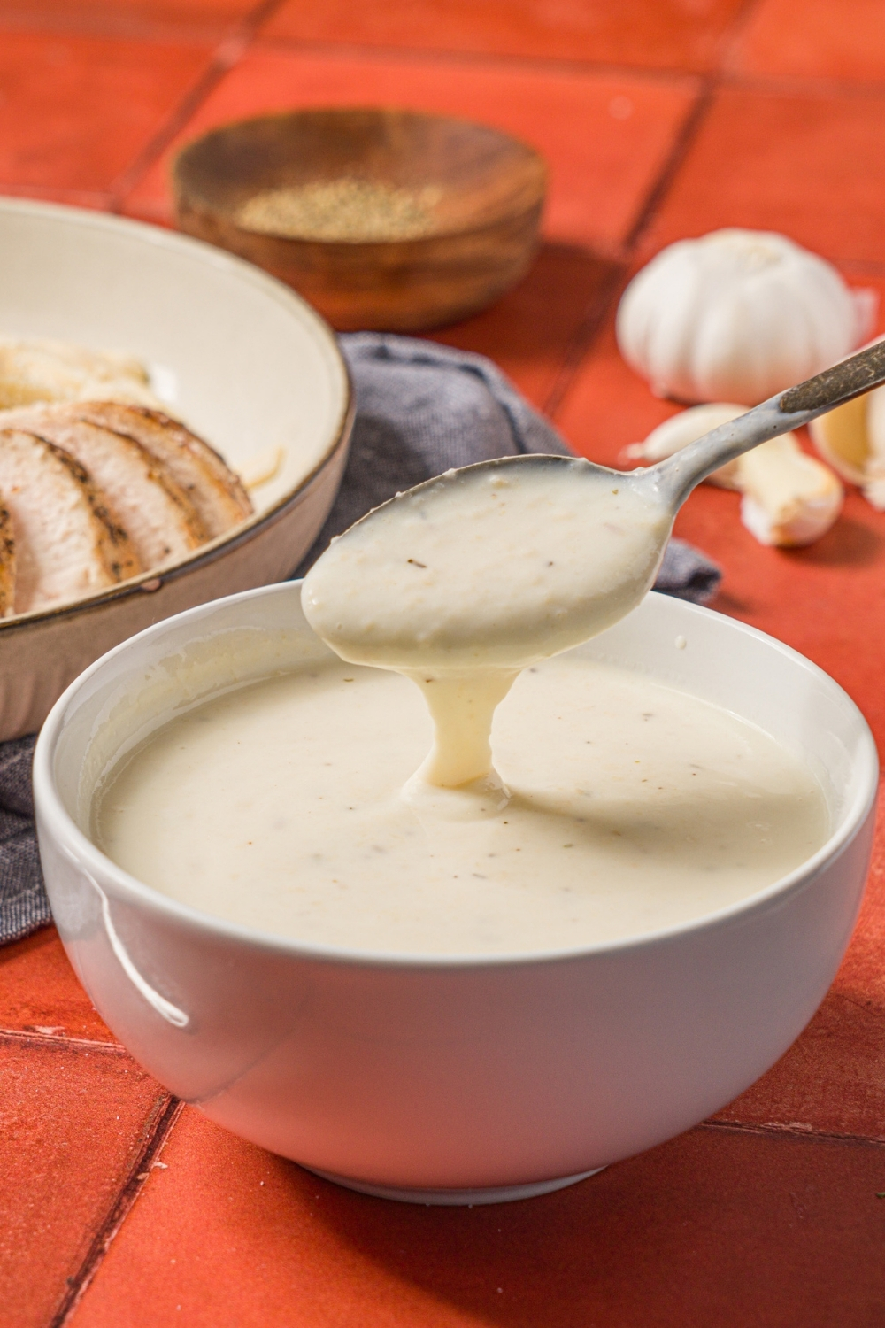 A bowl of alfredo sauce with a spoon dipping into the bowl. The bowl is on a brick counter with a bowl of chicken alfredo and small bowl of garnishes.