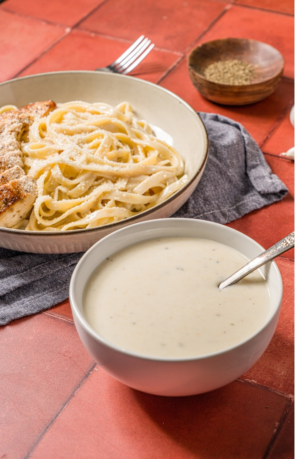 A bowl of alfredo sauce with a spoon in the bowl. The bowl is on a brick counter with a bowl of chicken alfredo and small bowl of garnishes.