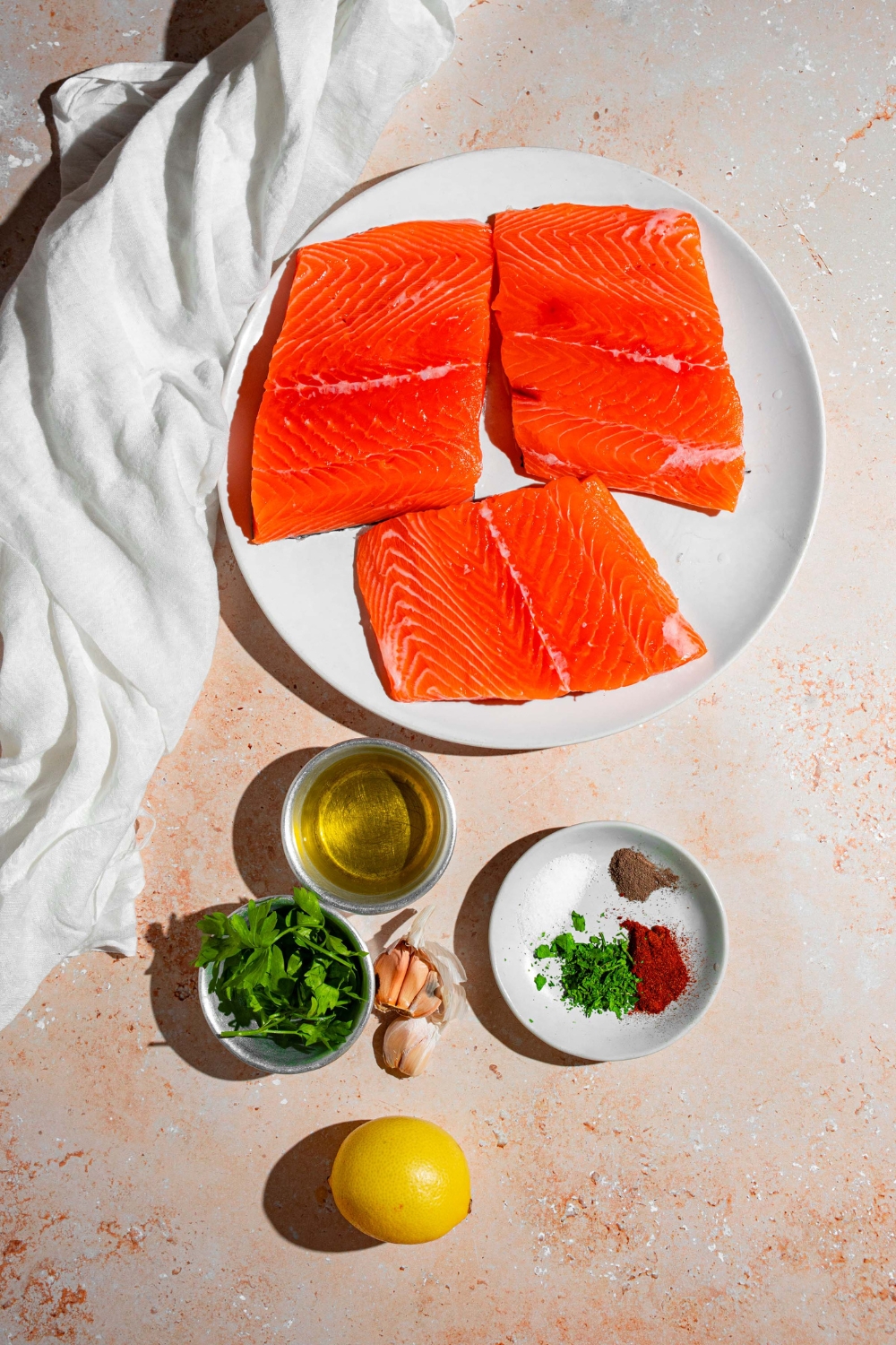 An overhead shot of several bowls in various sizes containing ingredients to make baked Sockeye salmon including Sockeye salmon, lemon, olive oil, garlic, parsley, and seasonings.