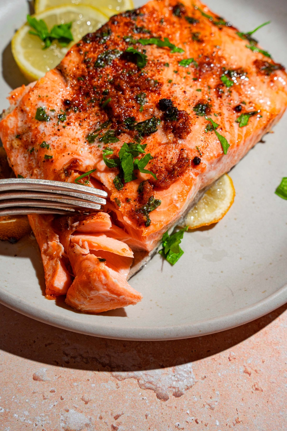 A white plate with baked Sockeye salmon garnished with fresh parsley and served with sliced lemon. There is a fork taking a bite from the salmon.
