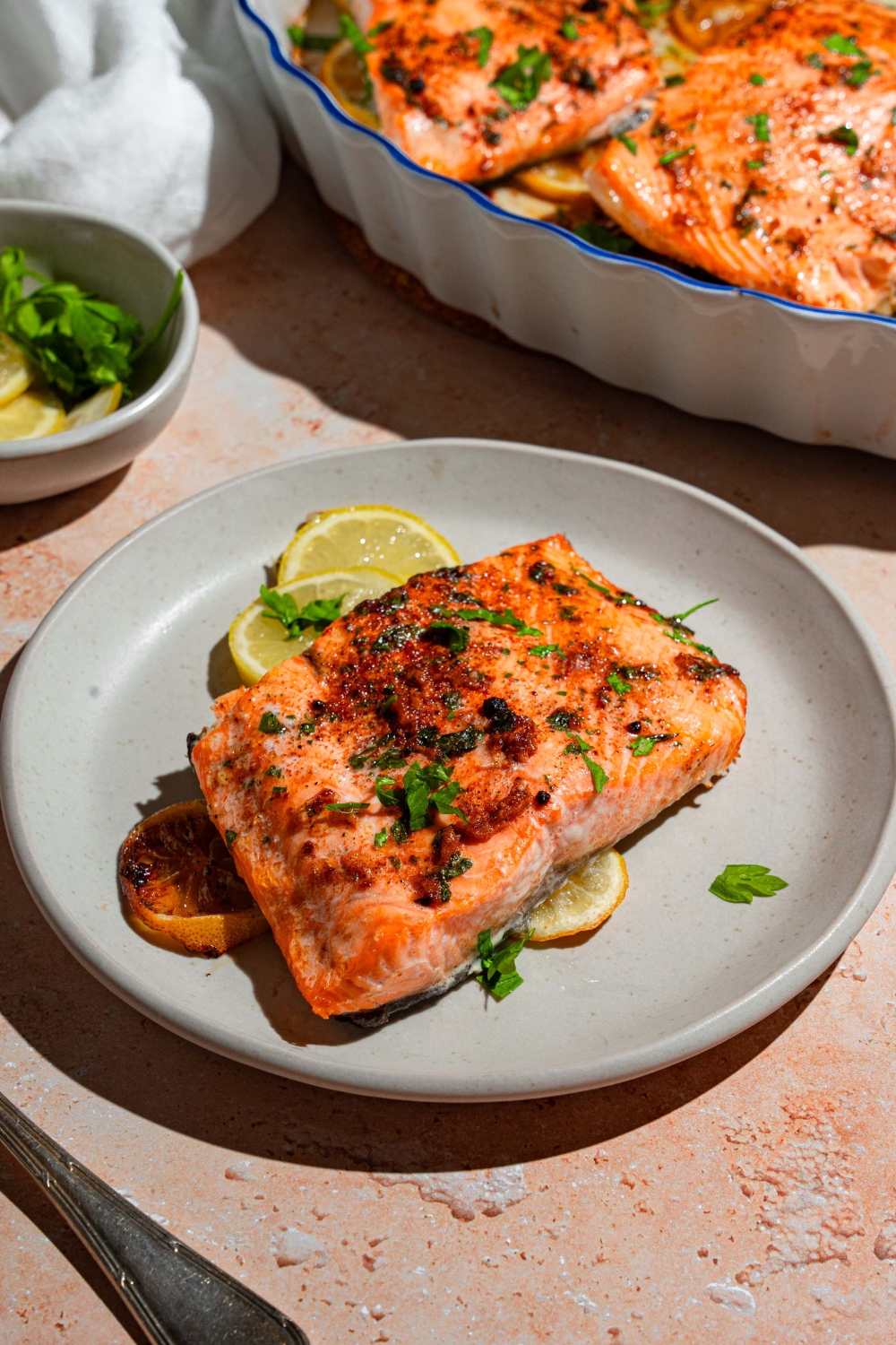A white plate with baked Sockeye salmon garnished with fresh parsley and served with sliced lemon. The plate is on a tan counter with a baking dish with salmon and small bowl of garnishes.