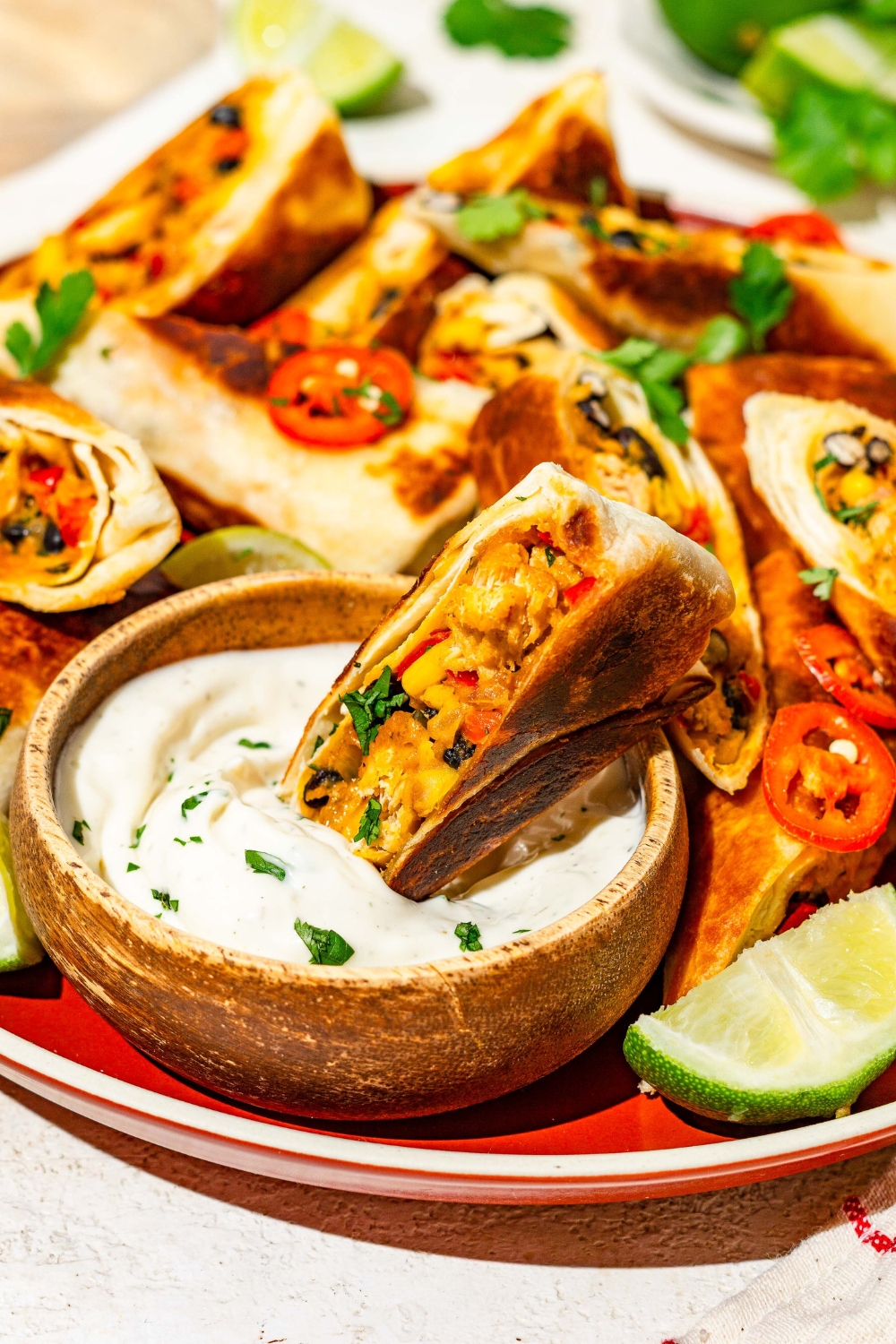 A plate of copycat Chili's southwestern egg rolls sliced in half and garnished with sliced peppers, fresh cilantro, and lime. There is a bowl of sour cream on the plate with an egg roll dipped in it.