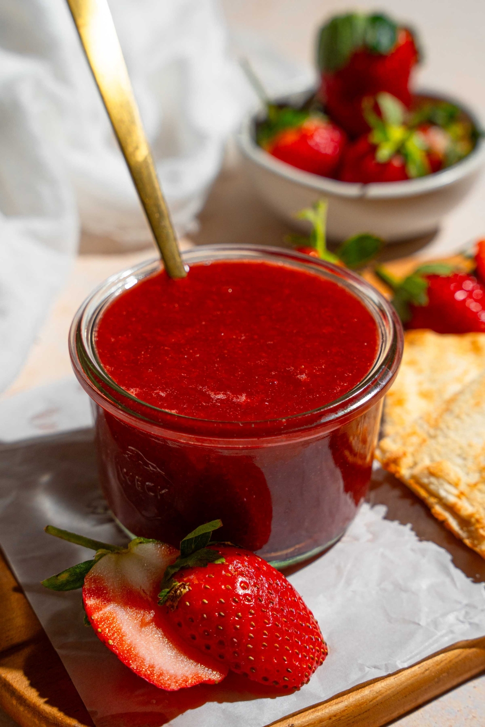 A jar of Christmas jam with a spoon dipped in jam. The jar is on a wooden board lined with parchment paper with a slice of toast and strawberries. The board is on a tan counter with a bowl of strawberries.