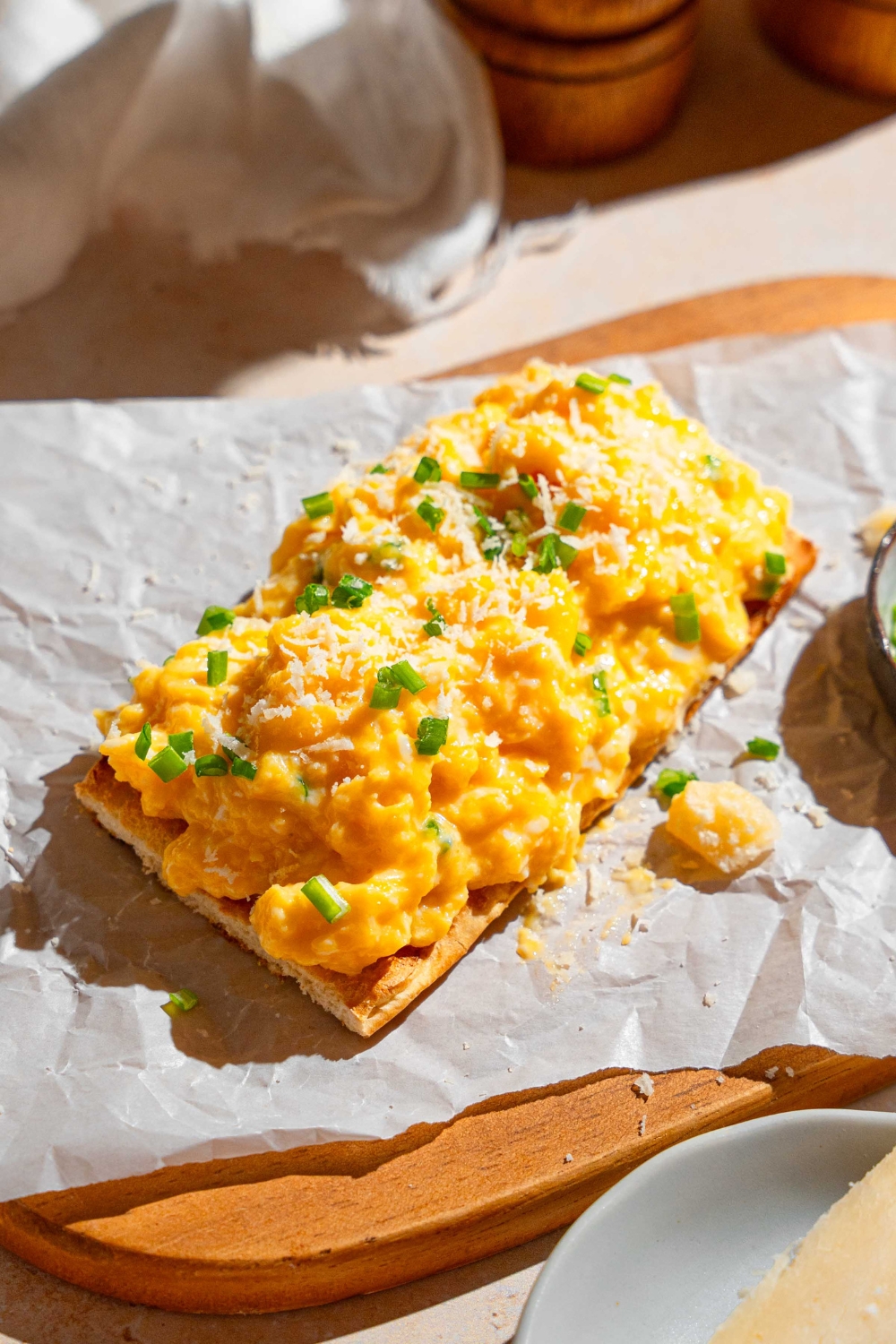 A thin slice toast topped with Gordon Ramsay scrambled eggs garnished with grated cheese and chives. The toast is on a wooden board lined with parchment paper and served with a ramekin of garnishes. The board is on a tan counter with a salt and pepper mill.