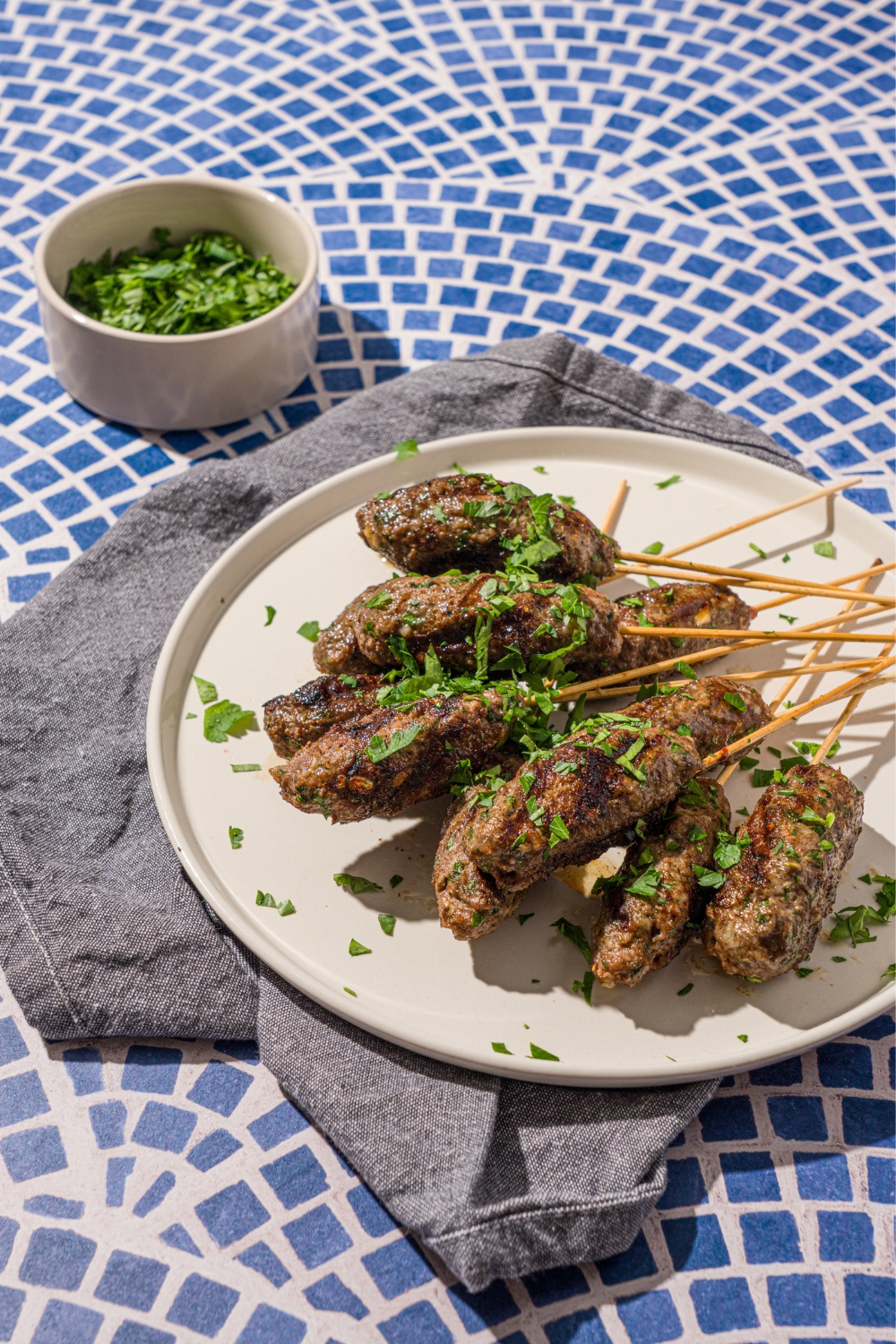A white plate with a pile of Kafta kabobs garnished with fresh parsley. The plate is on a tiled counter with a gray cloth napkin and small bowl of garnishes.