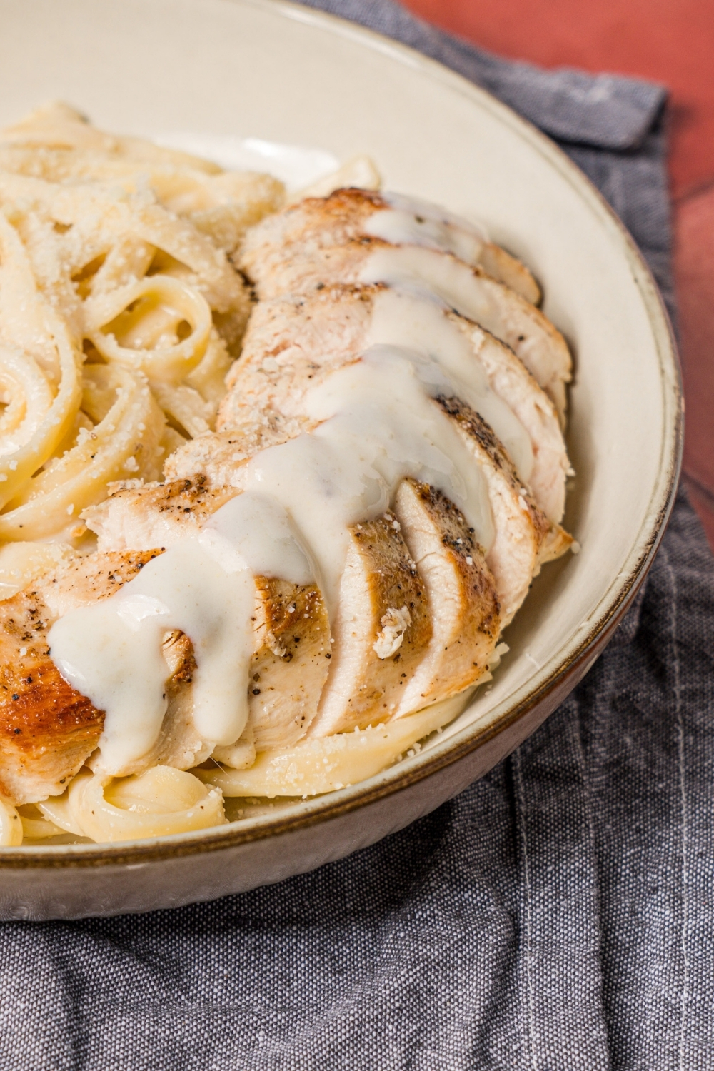 A bowl of chicken alfredo with alfredo sauce poured in a line over the chicken. The bowl is on a brick counter with a blue napkin.